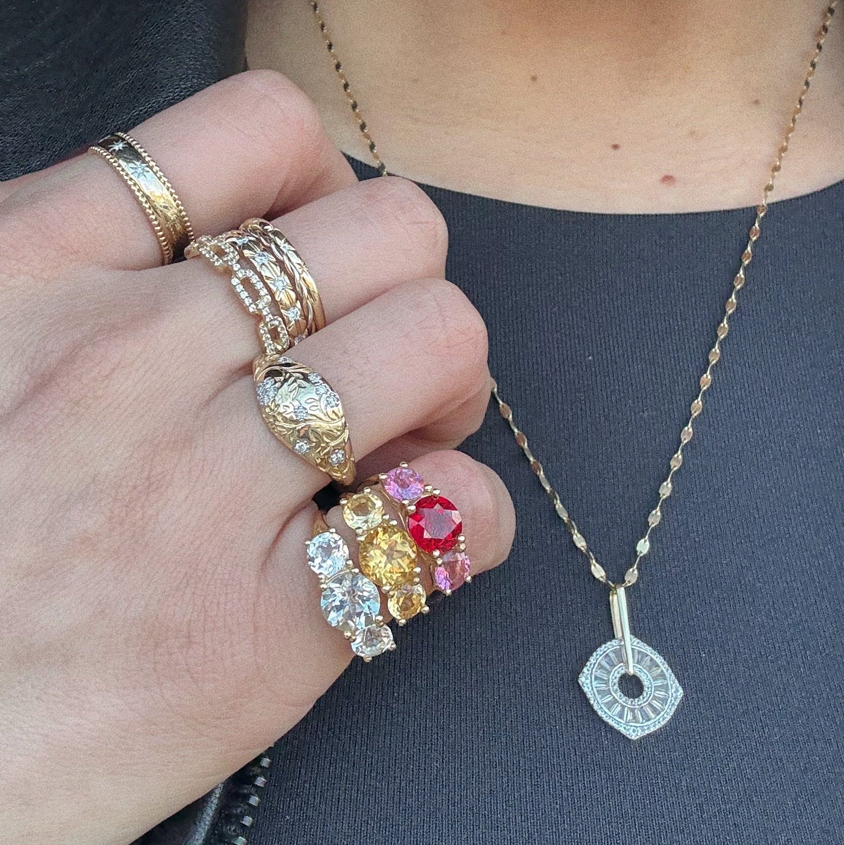 A close-up of a hand wearing the Solid Gold Trilogy Birthstone Pinky Ring Ruby Sample Size 4, paired with a 9k yellow gold chain necklace with a geometric pendant, set against a dark gray shirt.