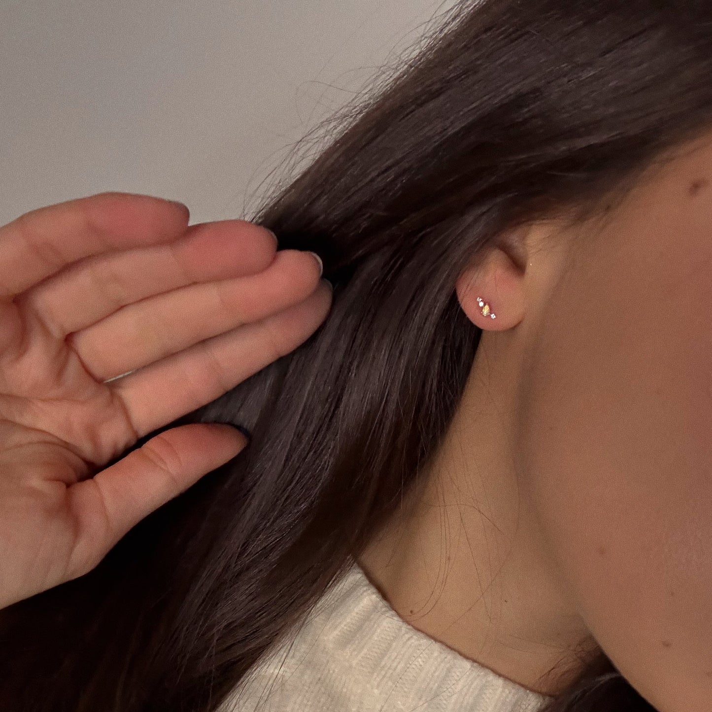 A person with long brown hair tucks it behind their ear, revealing a Solid Gold Opal Constellation Flat Back Stud. They're wearing a cream ribbed sweater against a simple neutral background.