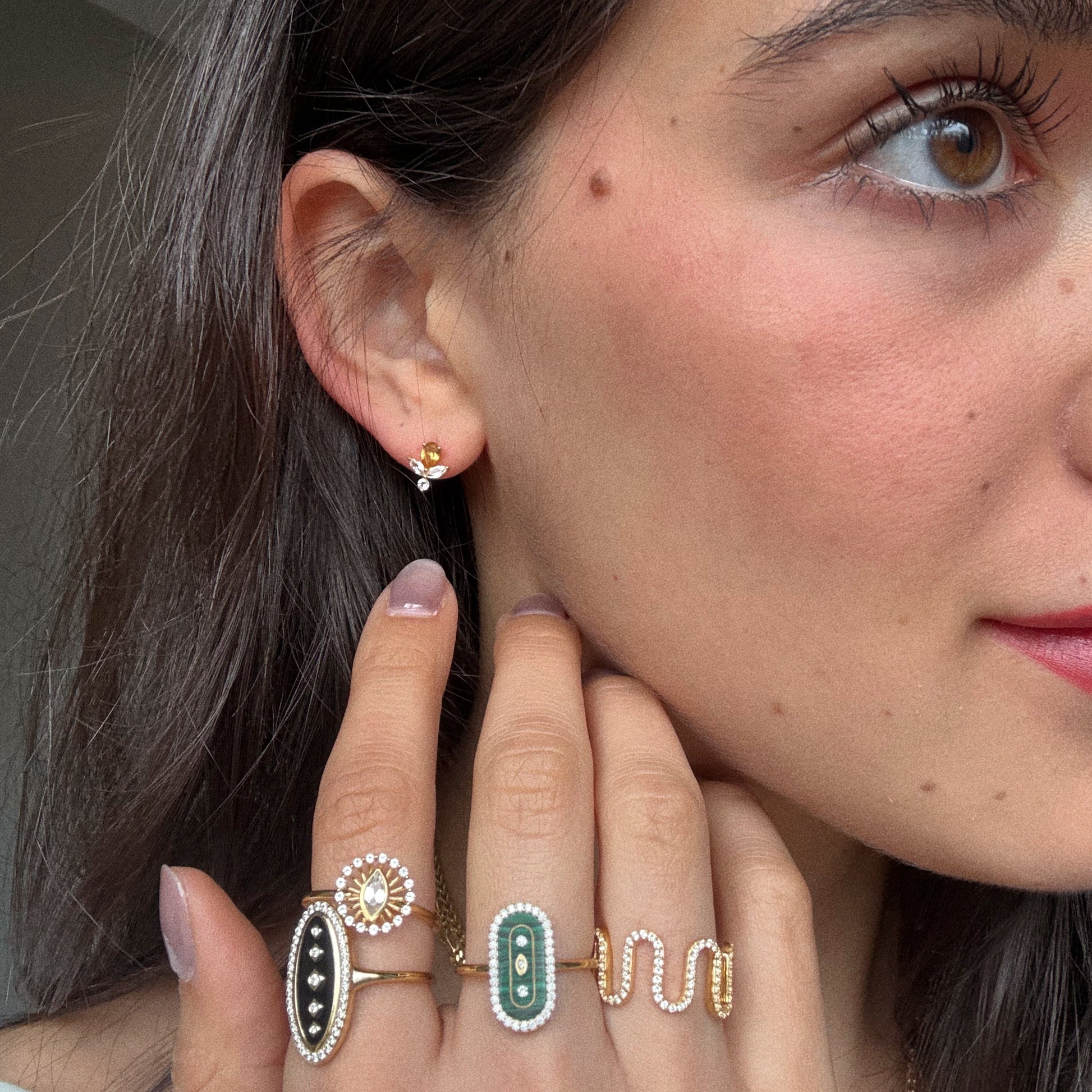 A close-up of a woman's face and hand highlights 14k yellow gold vermeil rings with green, white topaz, and black stones, along with the Jazz Age Citrine Studs floral gold earrings. Her nails are painted a soft mauve shade.