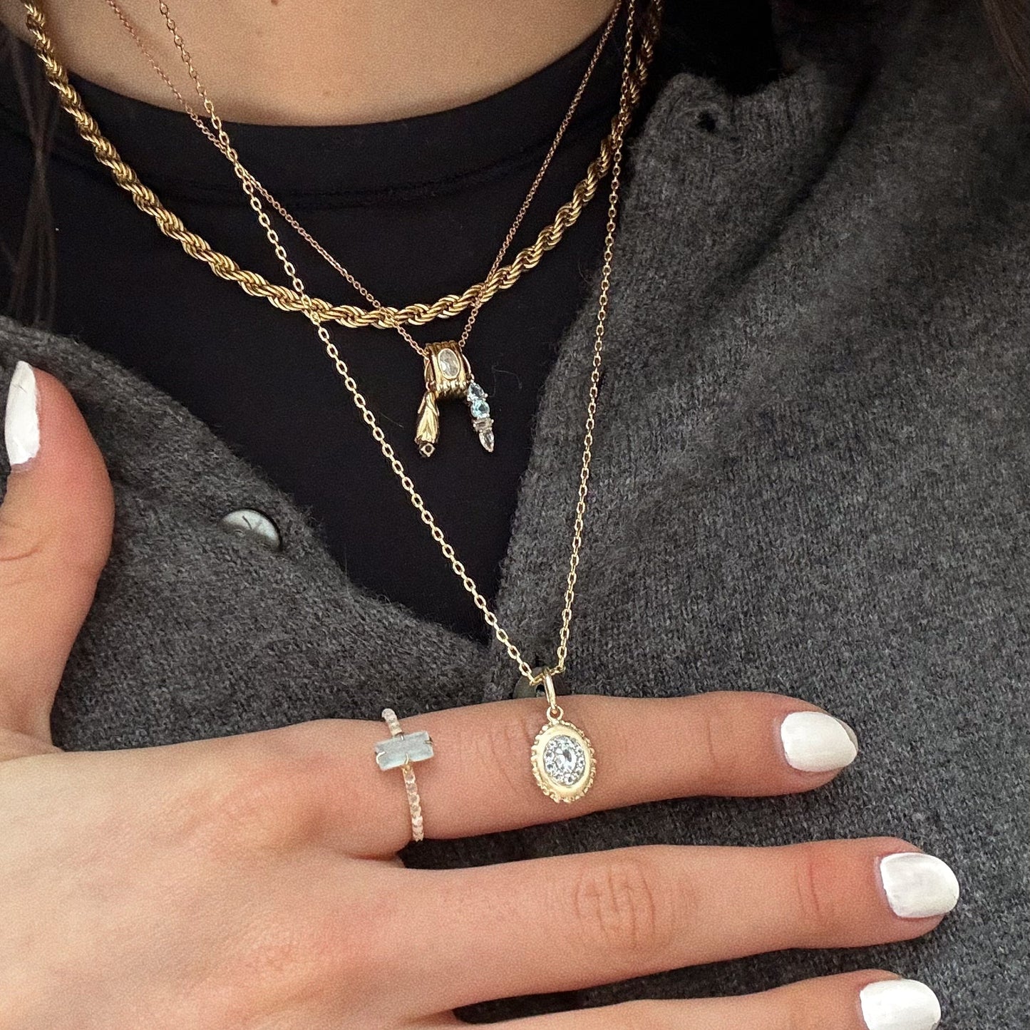 A close-up of a person wearing layered gold necklaces and the Solid Gold 2026 March Capsule Delicate Aquamarine Solitaire Ring, dressed in a gray buttoned shirt with white painted nails.