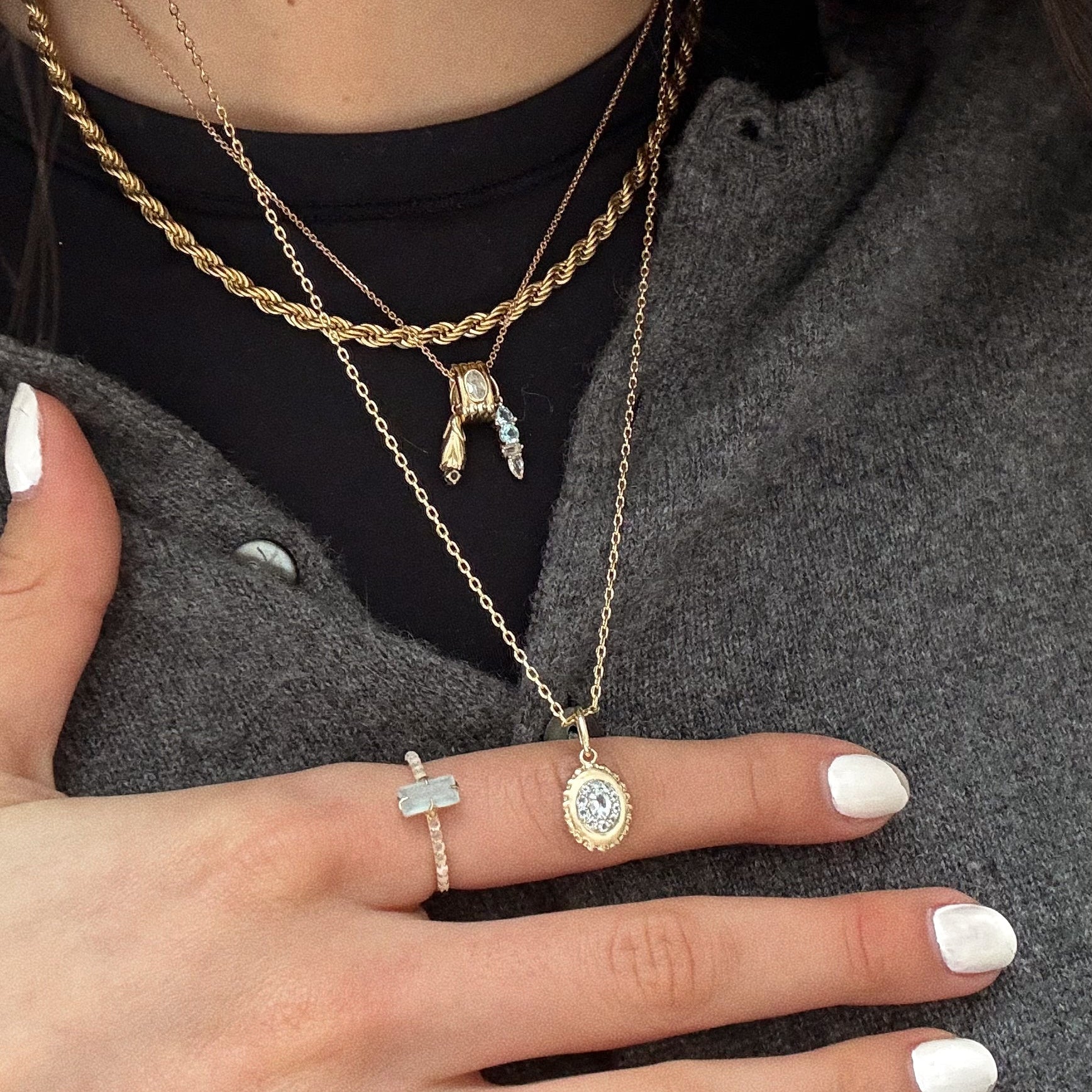 A close-up of a person wearing layered gold necklaces and the Solid Gold 2026 March Capsule Delicate Aquamarine Solitaire Ring, dressed in a gray buttoned shirt with white painted nails.