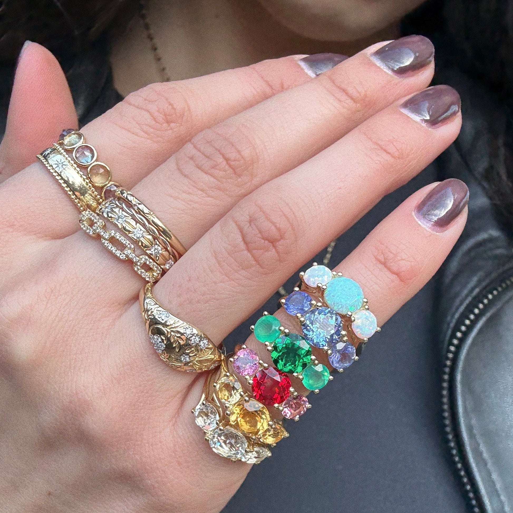 A hand with metallic silver nail polish wears the Solid Gold Trilogy Birthstone Pinky Ring in size 4, featuring 9k yellow gold, a 7 mm synthetic opal, diamonds, and colorful gemstones arranged in rainbow order on two fingers.