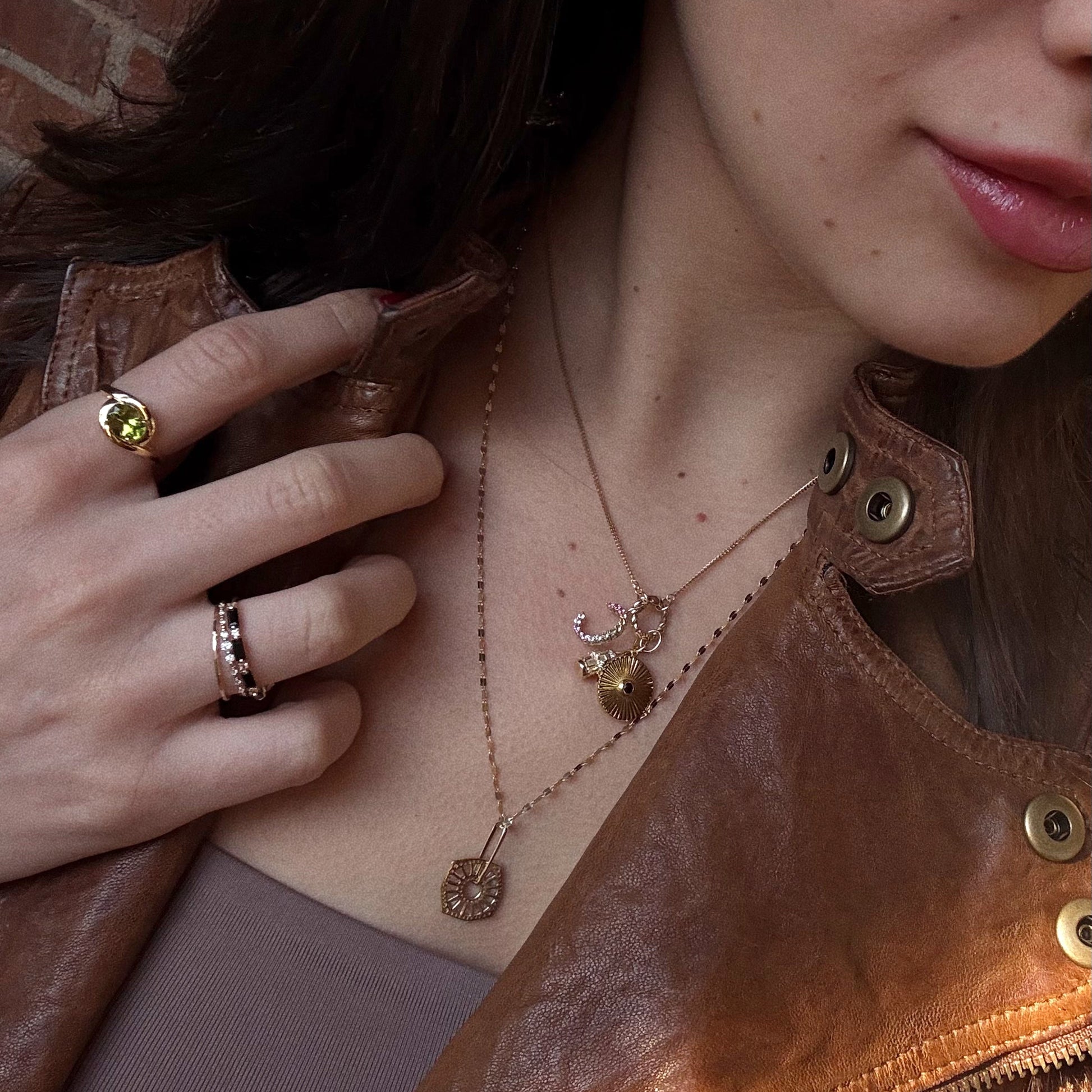 A woman in a brown leather jacket and taupe top showcases layered gold necklaces and the Peridot Poetry Statement Ring, a vintage-inspired 14k gold vermeil ring with a green gemstone, as she holds her jacket lapel near her face.