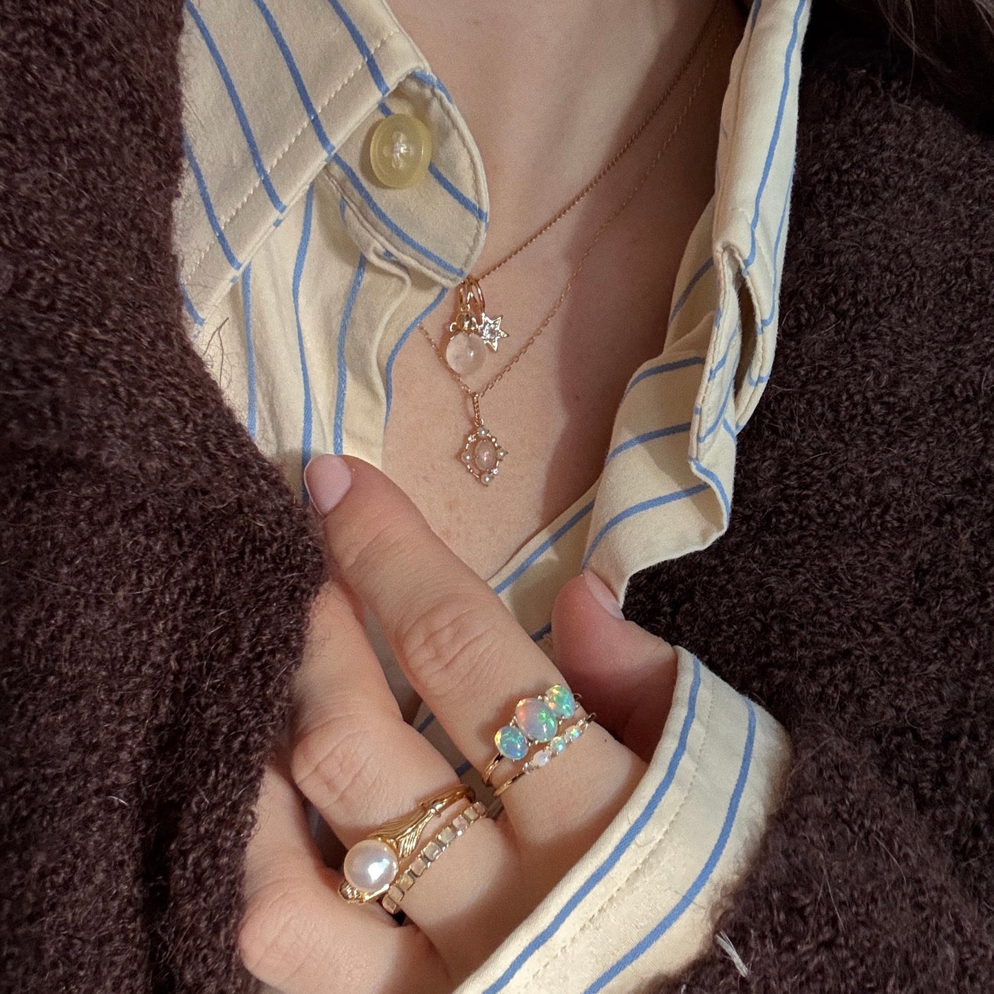A person wears gold rings, a Solid Gold Under The Sea Charm, and layered gold necklaces with pendants over a cream shirt with blue stripes and a dark brown textured jacket. Their nails are painted pale pink.