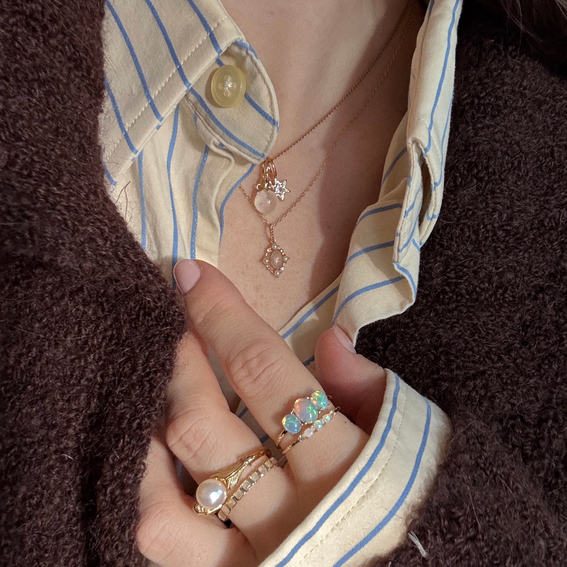 A person wears gold rings, a Solid Gold Under The Sea Charm, and layered gold necklaces with pendants over a cream shirt with blue stripes and a dark brown textured jacket. Their nails are painted pale pink.