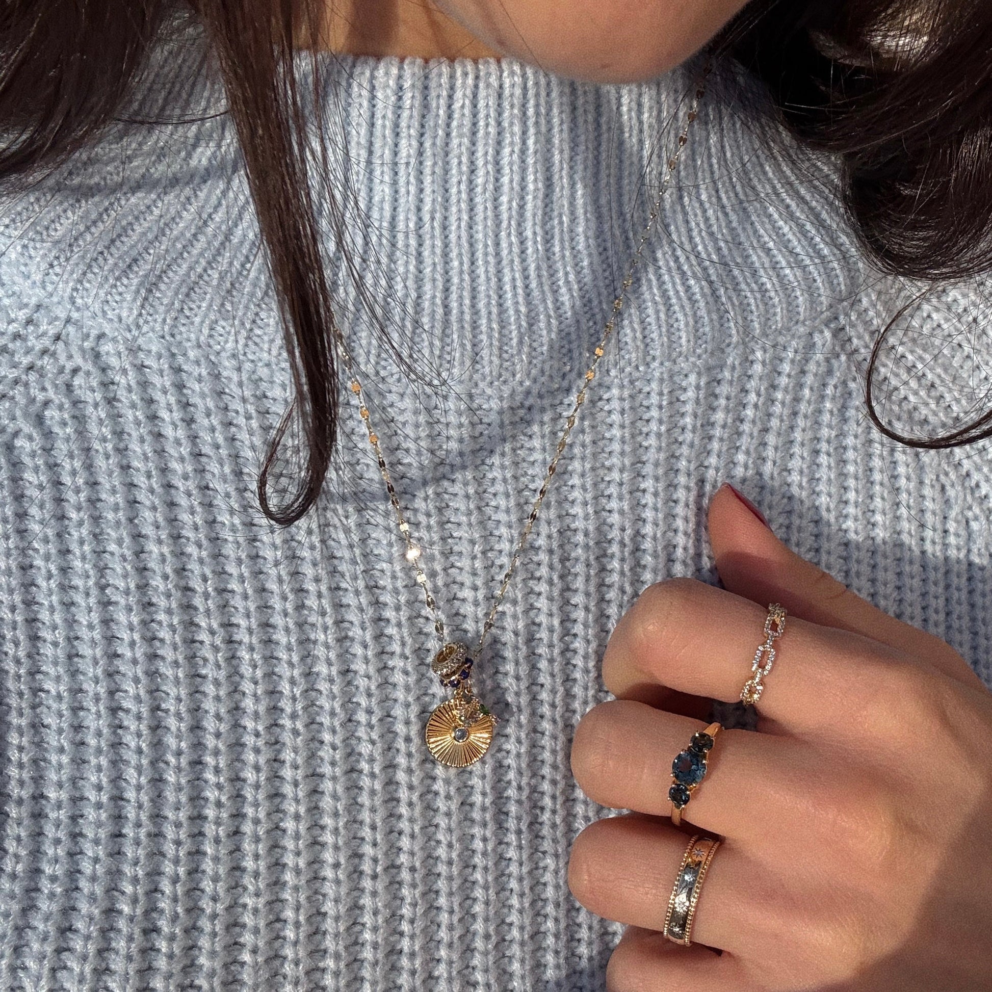 A person wearing a light blue knit sweater, a gold pendant necklace, and three rings—including the London Blue Topaz Trilogy Ring—holding their sweater near their chin.