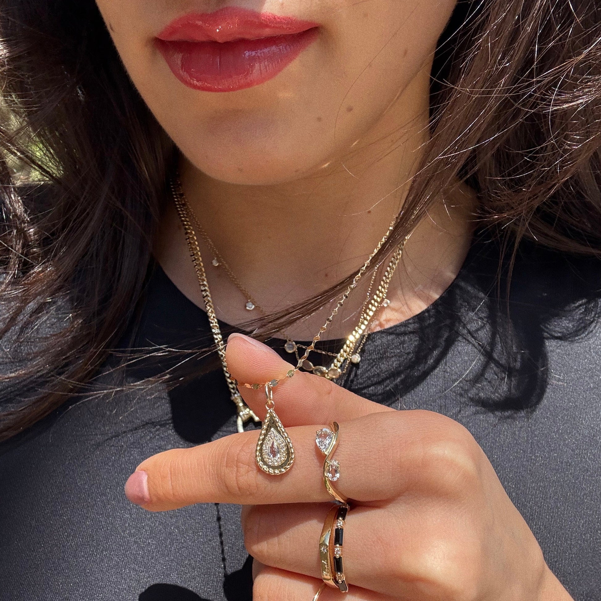 A woman with wavy dark hair and glossy red lips shows off layered yellow gold vermeil necklaces, including the 2026 April Capsule Tethered Teardrop White Topaz Charm, and two gold rings—all set against a black top.