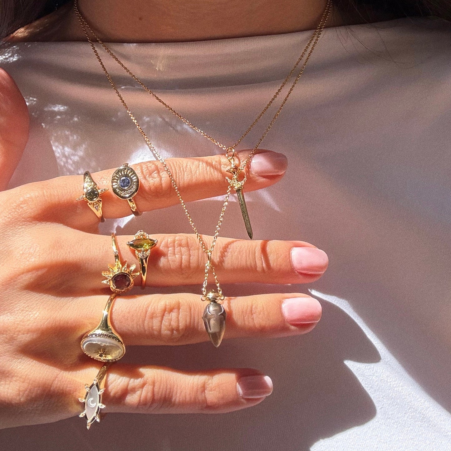 A manicured hand with multiple gold rings holds two layered gold necklaces, including the Buffy Dagger Necklace featuring a crescent moon dagger pendant, against a soft, light background.