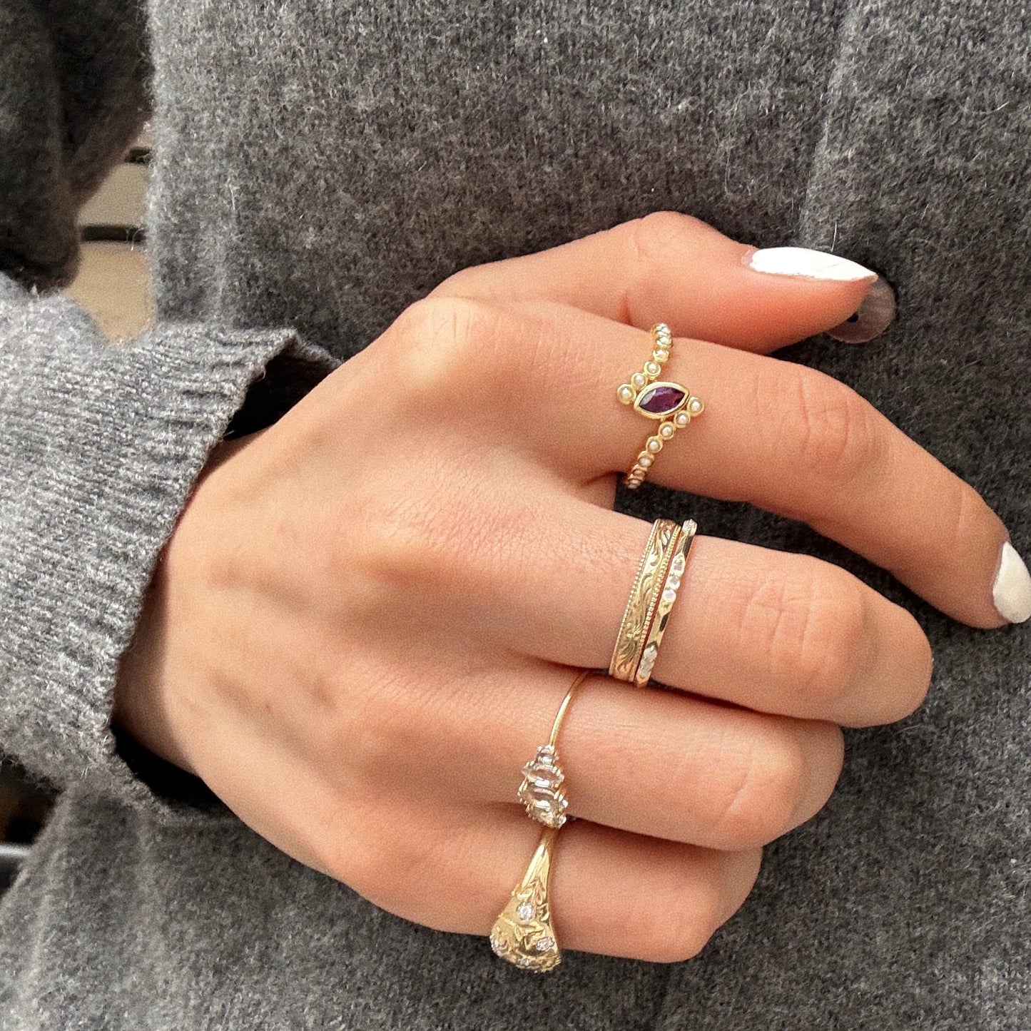 A close-up of a hand wearing the Solid Gold 2026 February Capsule Floating Amethyst and Pearl Twist Ring with intricate gold bands, against a gray wool sweater. The nails are neatly manicured and painted white; background softly blurred.