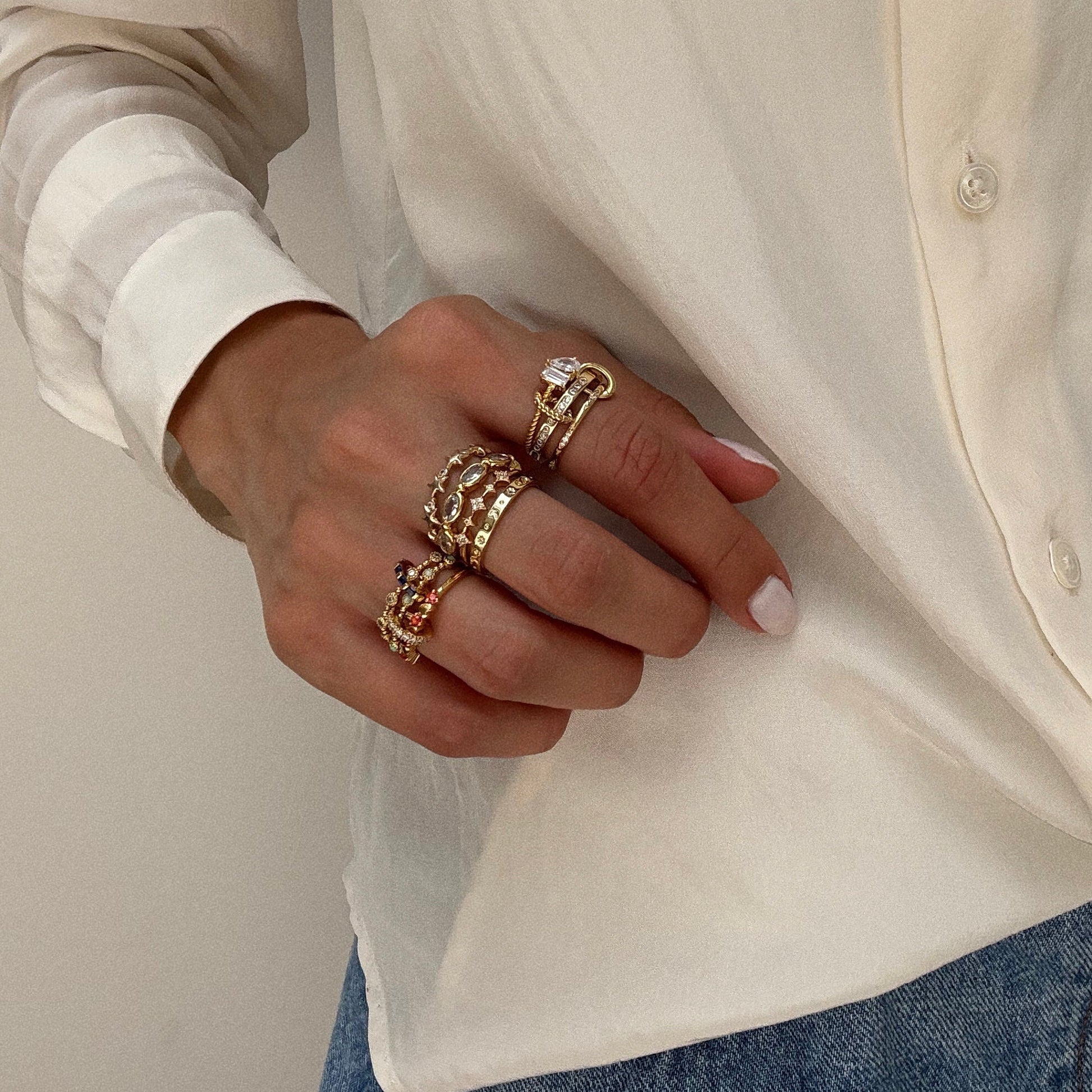A hand with a natural manicure wears multiple chunky gold rings and the Ring Clip from Local Eclectic, shown against a cream-colored blouse and blue jeans.