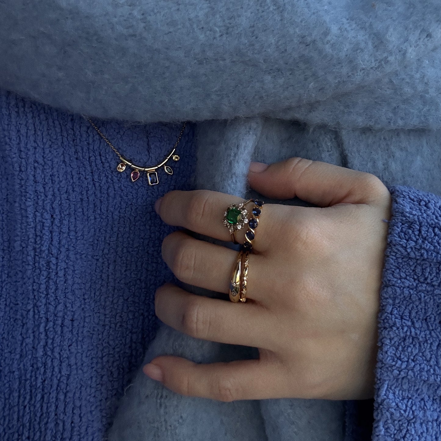 A hand wearing the Green Gardens Vintage Inspired Ring rests on soft, textured light blue fabric, with a gold necklace featuring colorful stone pendants visible near the collar.