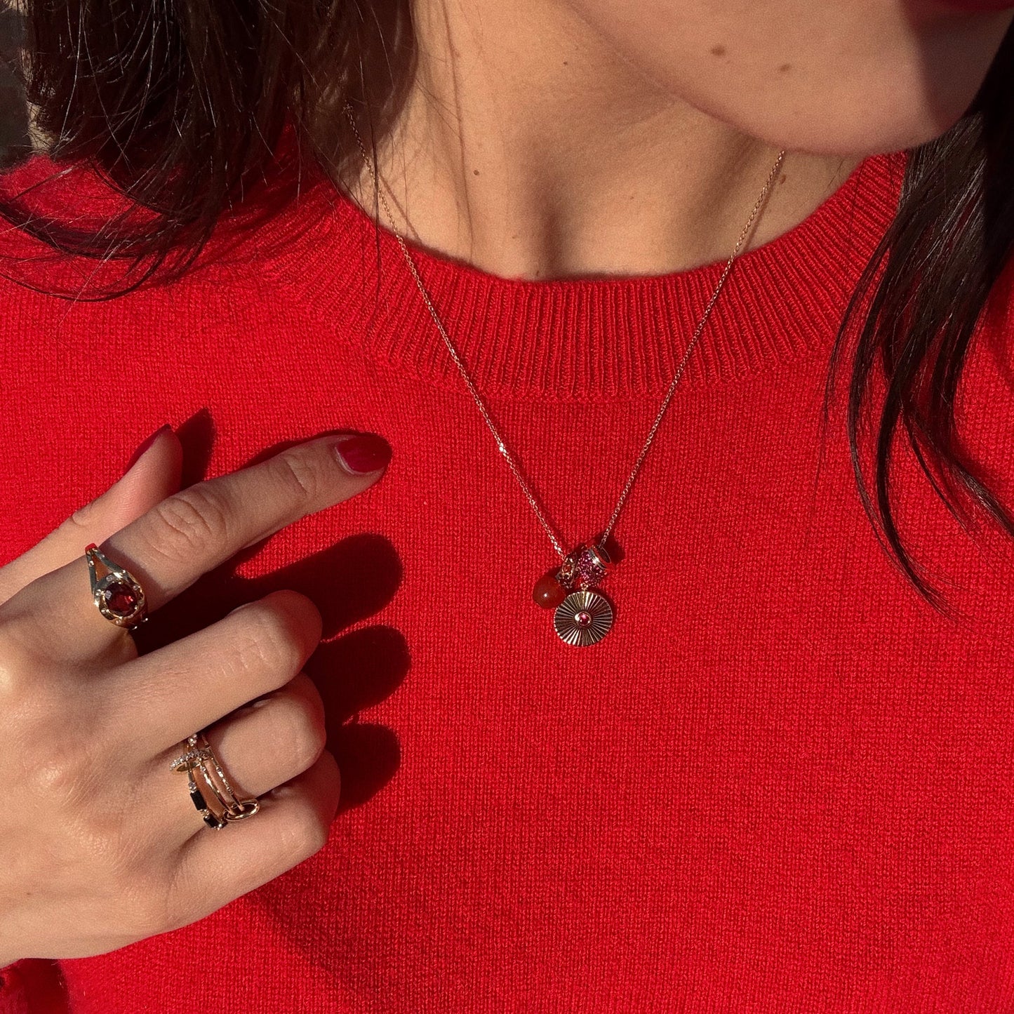 A woman in a bright red sweater models rings, including the striking Golden Age Garnet Cocktail Ring, with red nails for a bold look. She also wears a gold necklace adorned with small charms.