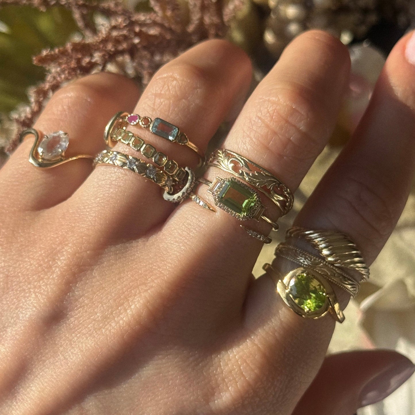 A hand displays multiple gold rings, including the 14k Open Front Stacking Ring crafted from recycled gold, alongside bands with green gemstones and white sapphire, all featuring intricate designs and sparkling in natural light.