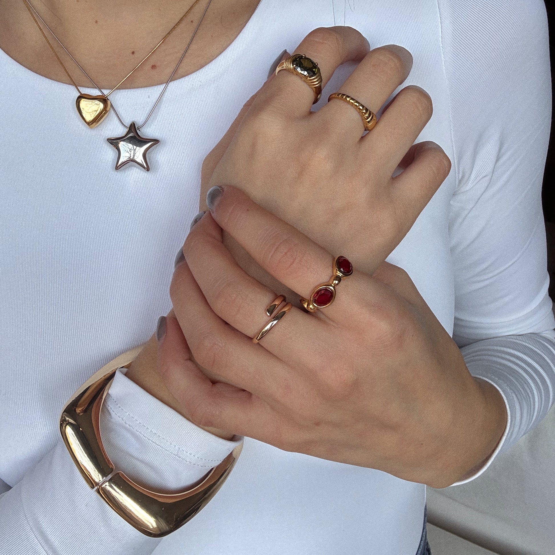 A person in a white long-sleeve top shows off layered gold necklaces, gold rings with red stones, the Square Cuff Bracelet, and light blue nail polish on crossed hands.