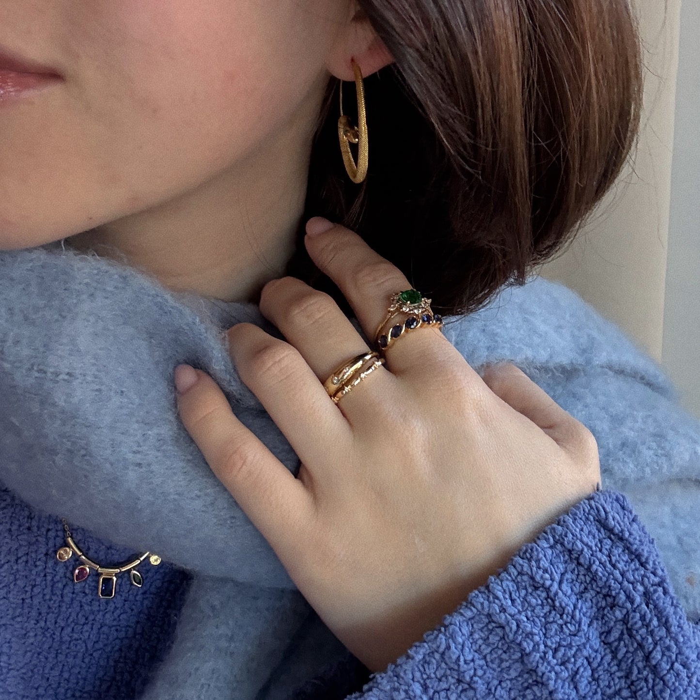 A woman in gold hoop earrings, gemstone rings, and a vibrant necklace styles her blue scarf and sweater—an inspiring holiday look that's perfect for gifting or unboxing the 2025 Jewelry Advent Calendar this season.