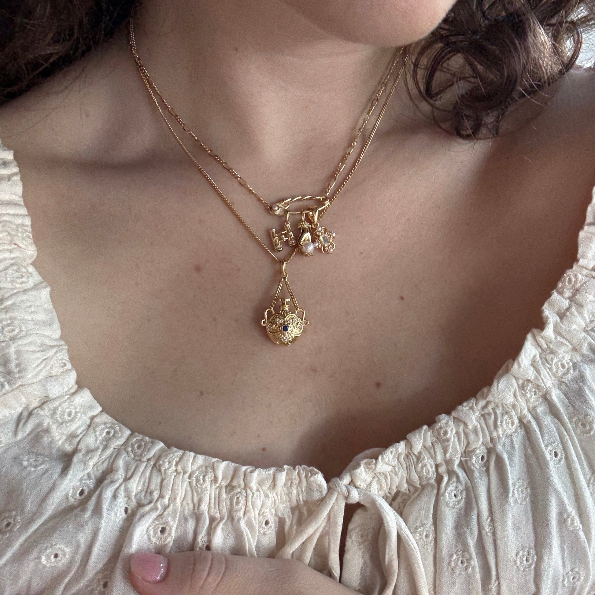 A woman wears layered gold necklaces, featuring the Vintage Inspired Pendant Charm and pearls, over a cream embroidered blouse with a gathered neckline.