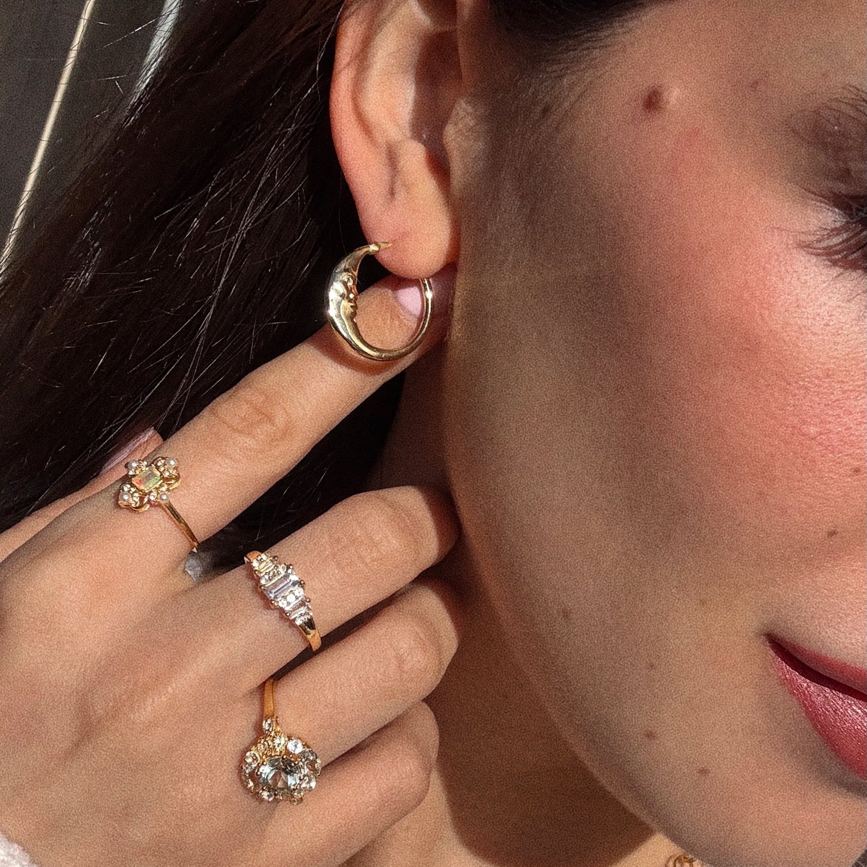 A close-up of a woman’s face and hand highlights her wearing Petite Luna Hoops, yellow gold plated earrings, along with several gold rings featuring flower and diamond designs. She touches the earring, showing off her natural makeup look.