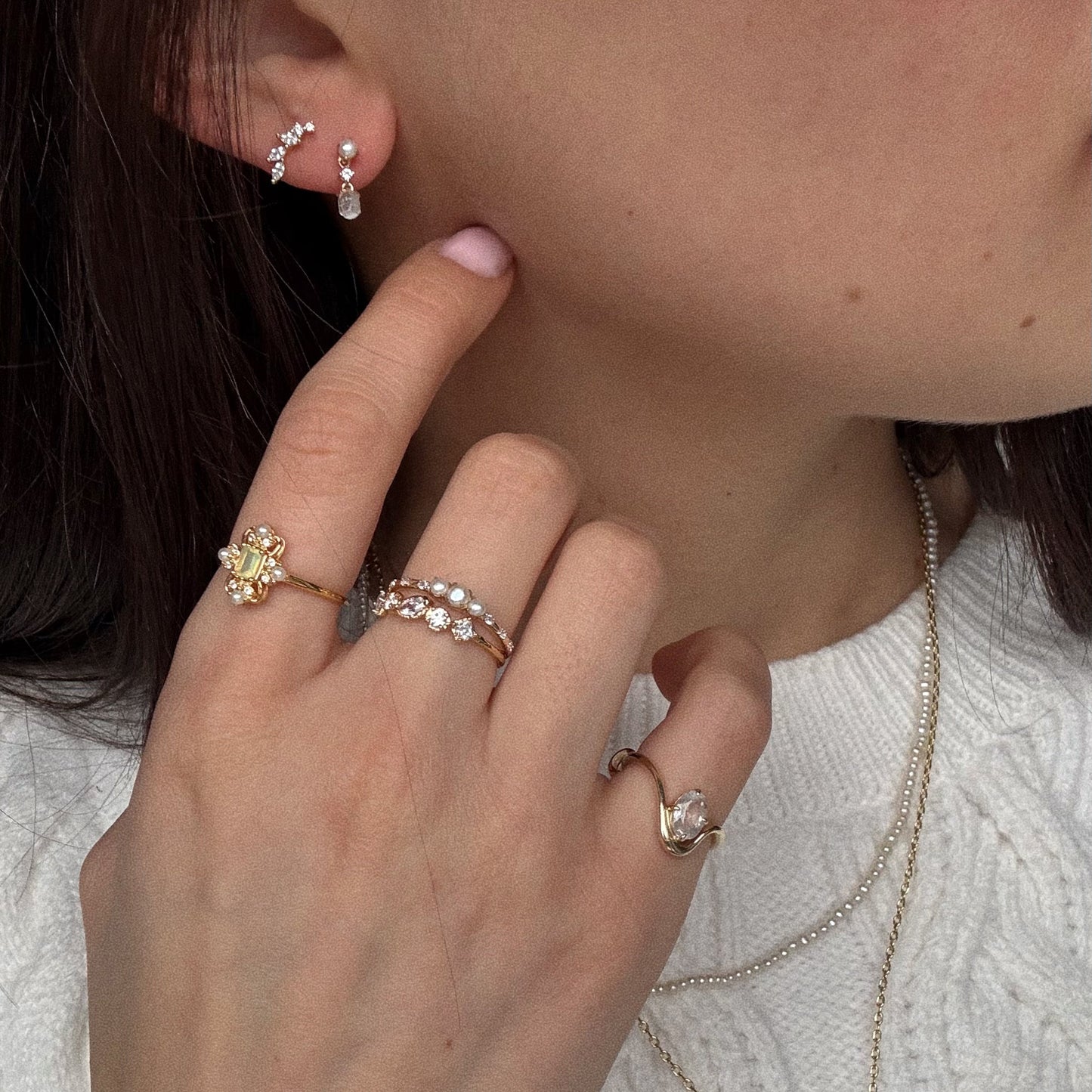 A woman in a white sweater shows her hand near her face, wearing the Solid Gold Luminous Pearl Trio Ring, layered necklaces, and pearl-crystal earrings. Her short fingernails are painted pale pink.