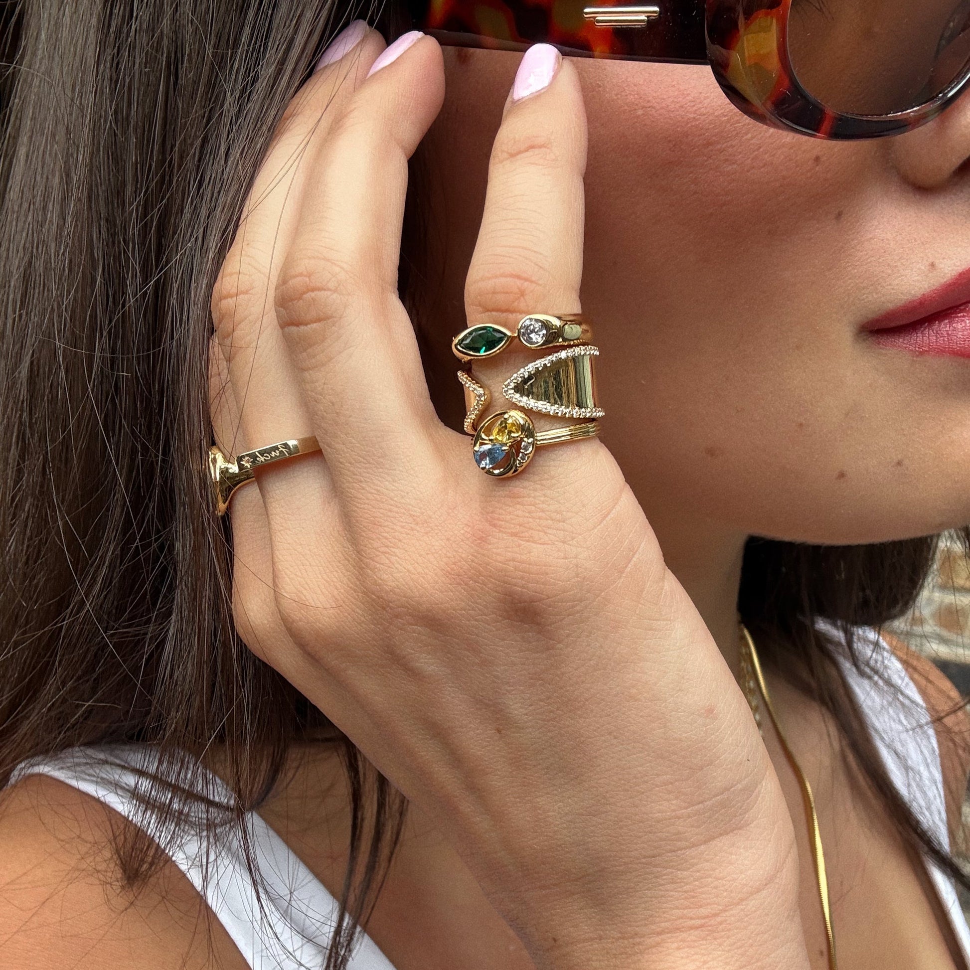 A woman wearing sunglasses touches her face, displaying multiple 14k yellow gold plated rings—including the Open Front Cigar Pinky Ring with a CZ border—alongside a simple gold necklace and light pink nails.
