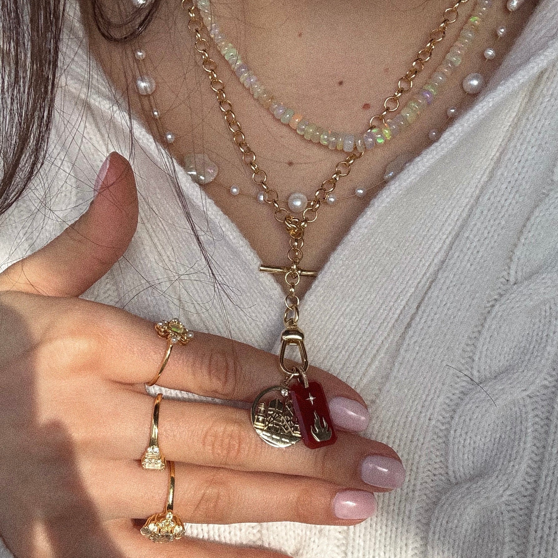 A close-up of a person wearing layered gold and beaded necklaces, including the Solid Gold Crystal Element Charm, gold rings with floral designs, and a cream cable-knit sweater. Their nails are polished pale pink.