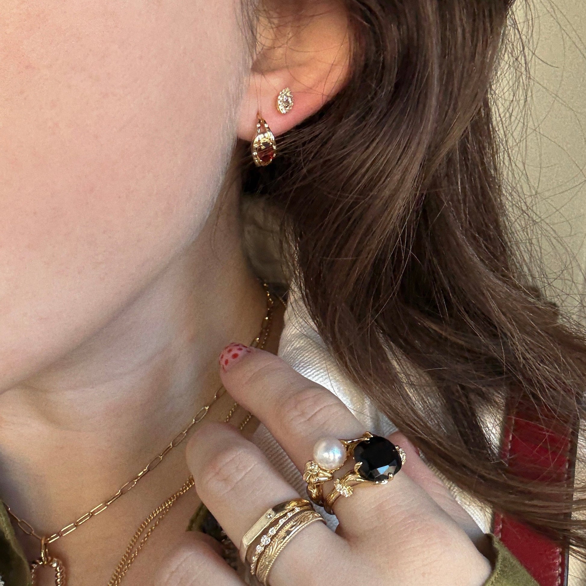 A close-up of a woman's ear and hand featuring Gilded Garnet Huggie Hoops, gemstone necklaces and rings, with wavy brown hair, patterned pink nails, and a collared shirt.