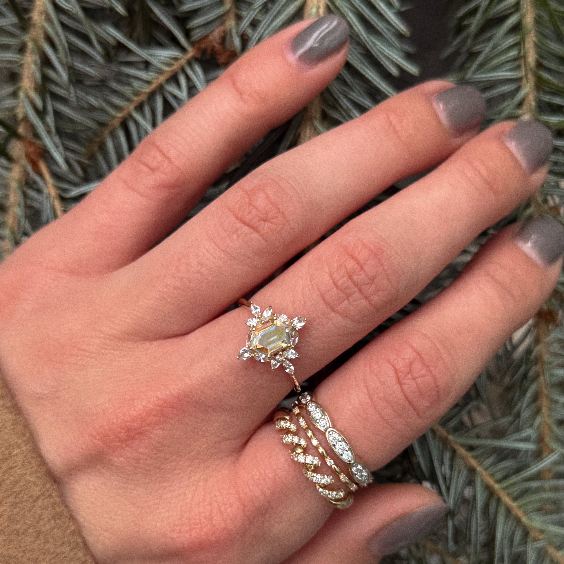 A hand with grey-painted nails showcases the romantic 14k Fated Twist Diamond Ring and two 14k solid yellow gold bands, all set against pine tree branches.