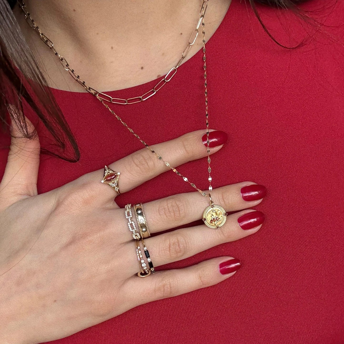 A person with red nail polish and multiple gold rings and necklaces, including the 2026 January Capsule Vintage Inspired Garnet Coin Charm, rests their hand on their chest over a red top.