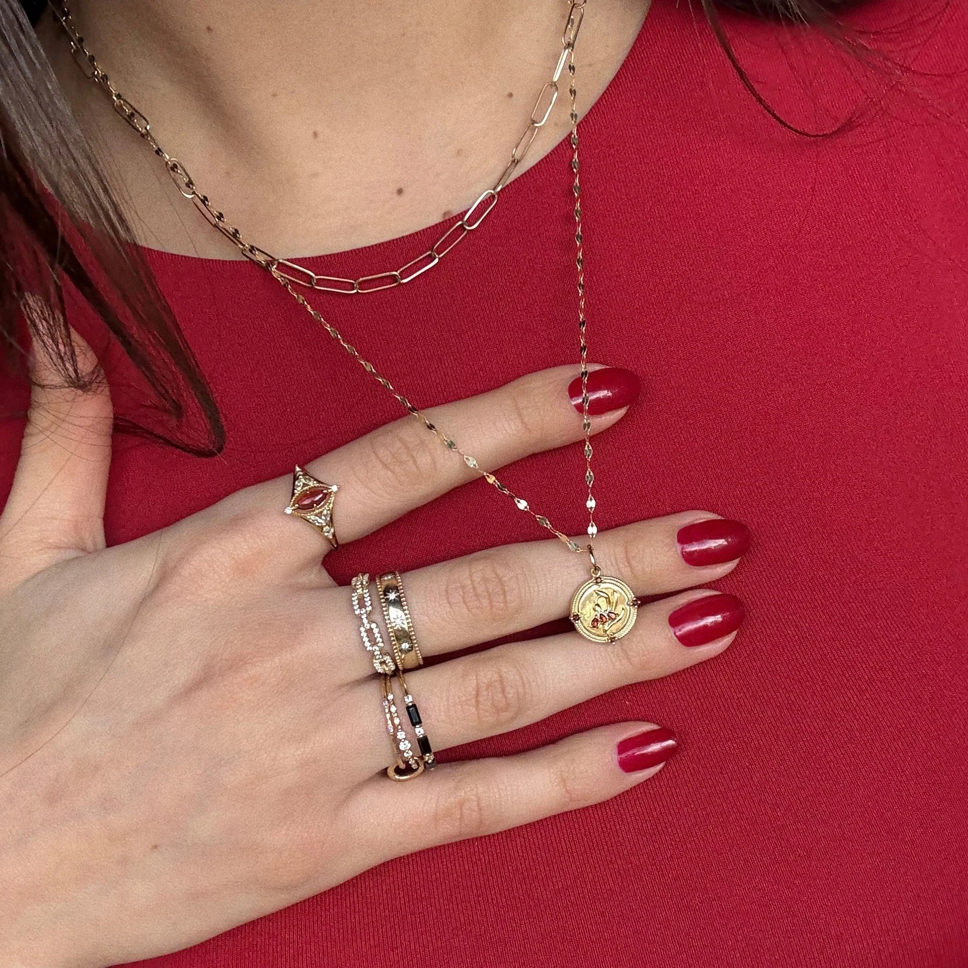 A person with red nail polish and multiple gold rings and necklaces, including the 2026 January Capsule Vintage Inspired Garnet Coin Charm, rests their hand on their chest over a red top.
