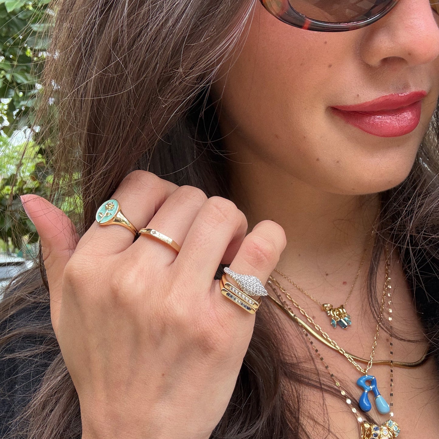 A woman with long brown hair and red lipstick wears sunglasses, several gold rings—including the Pave Signet Pinky Ring—layered gold necklaces, and a black top. She poses outdoors, holding her hand near her chin to show off her jewelry.