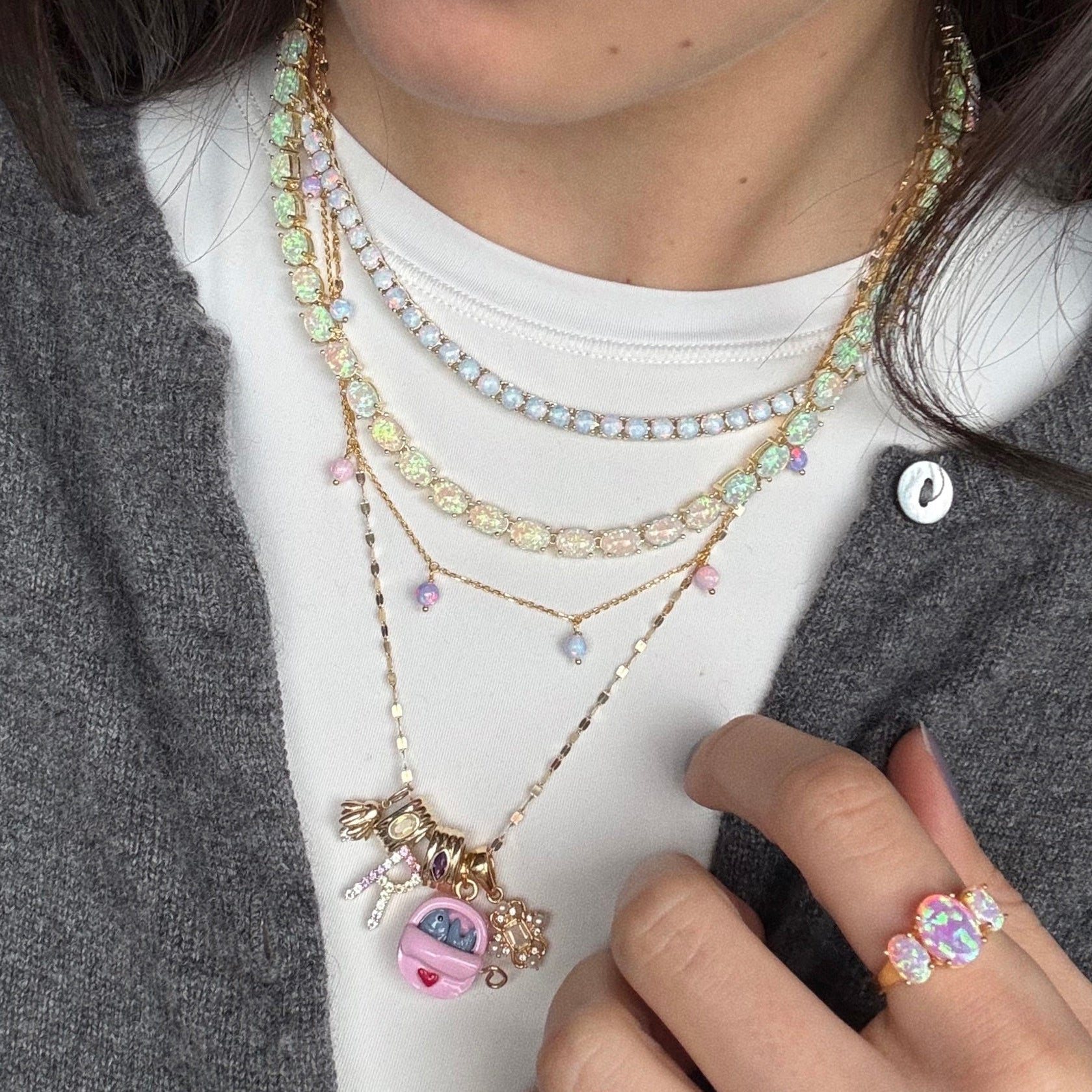 A person wears layered pastel beaded necklaces, including the Solid Gold Bold Birthstone Spacer Charm and a charm necklace with a pink Tamagotchi. They also have a pastel beaded ring and wear a grey cardigan over a white top.