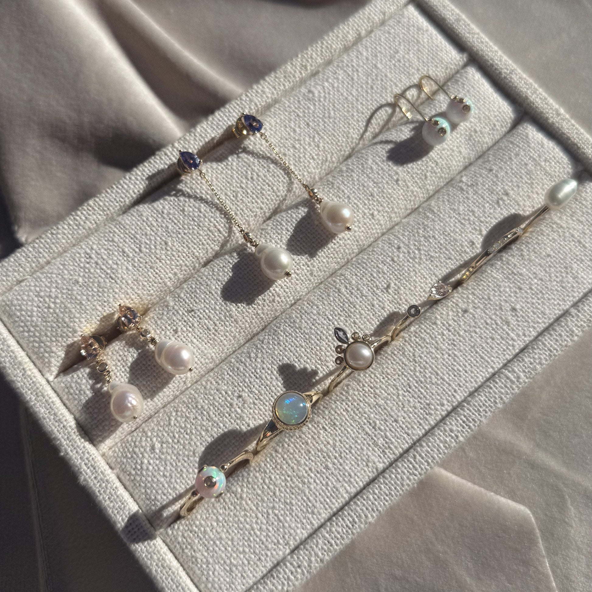 A jewelry box displays two pairs of Solid Gold Floating Opal Gem Drop Earrings (17 mm long) and a delicate gold bracelet with pearls, opals, and decorative accents, all set on light beige fabric.