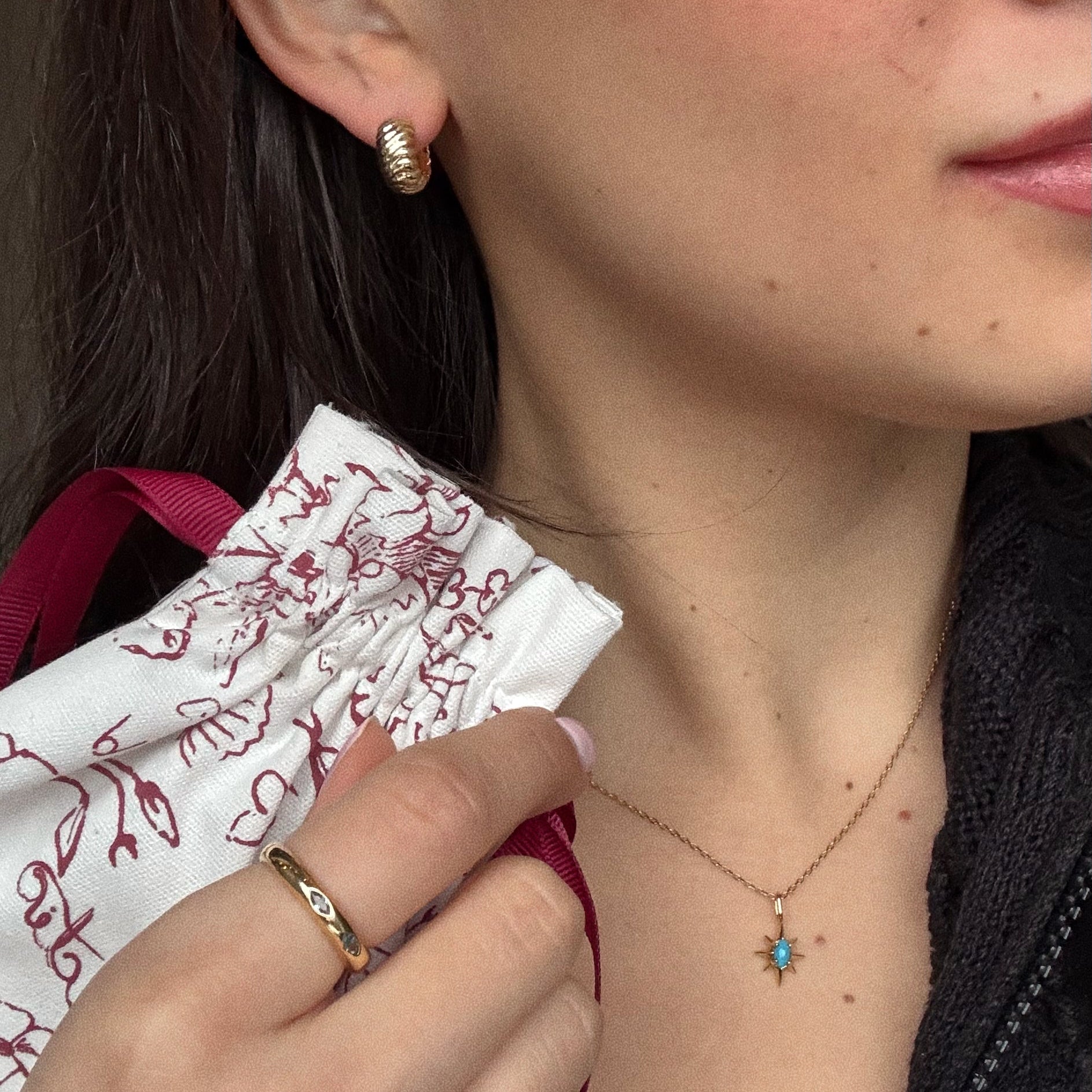A woman wears gold hoop earrings, a gold ring, and a gold necklace with a blue star pendant. She holds the Surprise Box!—a white drawstring bag with red floral patterns—near her neck. Only her lower face and hand are visible.