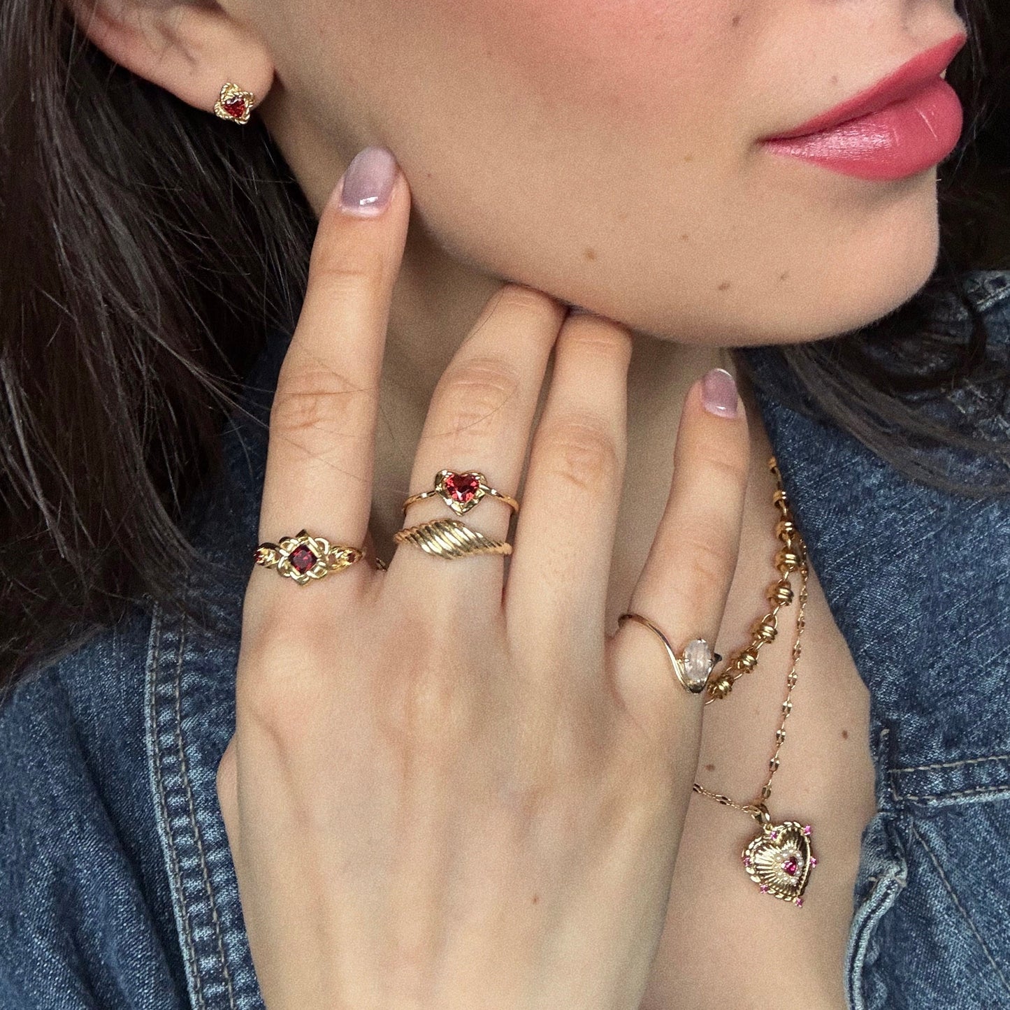 A woman in a denim jacket, pink lipstick, and manicured nails wears the Lover's Knot Heart Ring and matching 14k yellow gold vermeil jewelry with red heart-shaped stones—bracelet, earrings, and necklace.