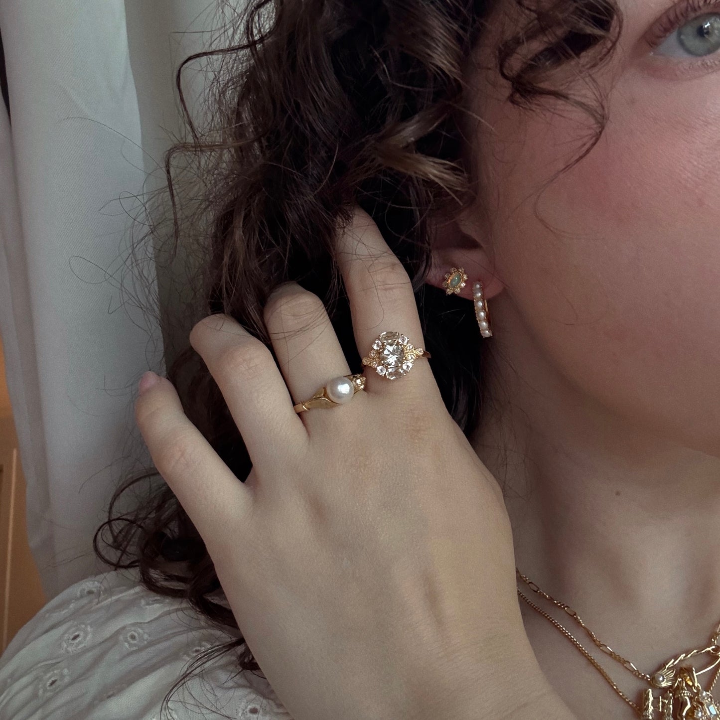 A close-up of a person with curly brown hair wearing vintage gold jewelry, featuring the Opal Majesty Studs, pearl and gem rings, layered necklaces, and gold earrings. They touch their face, partially covering one eye.