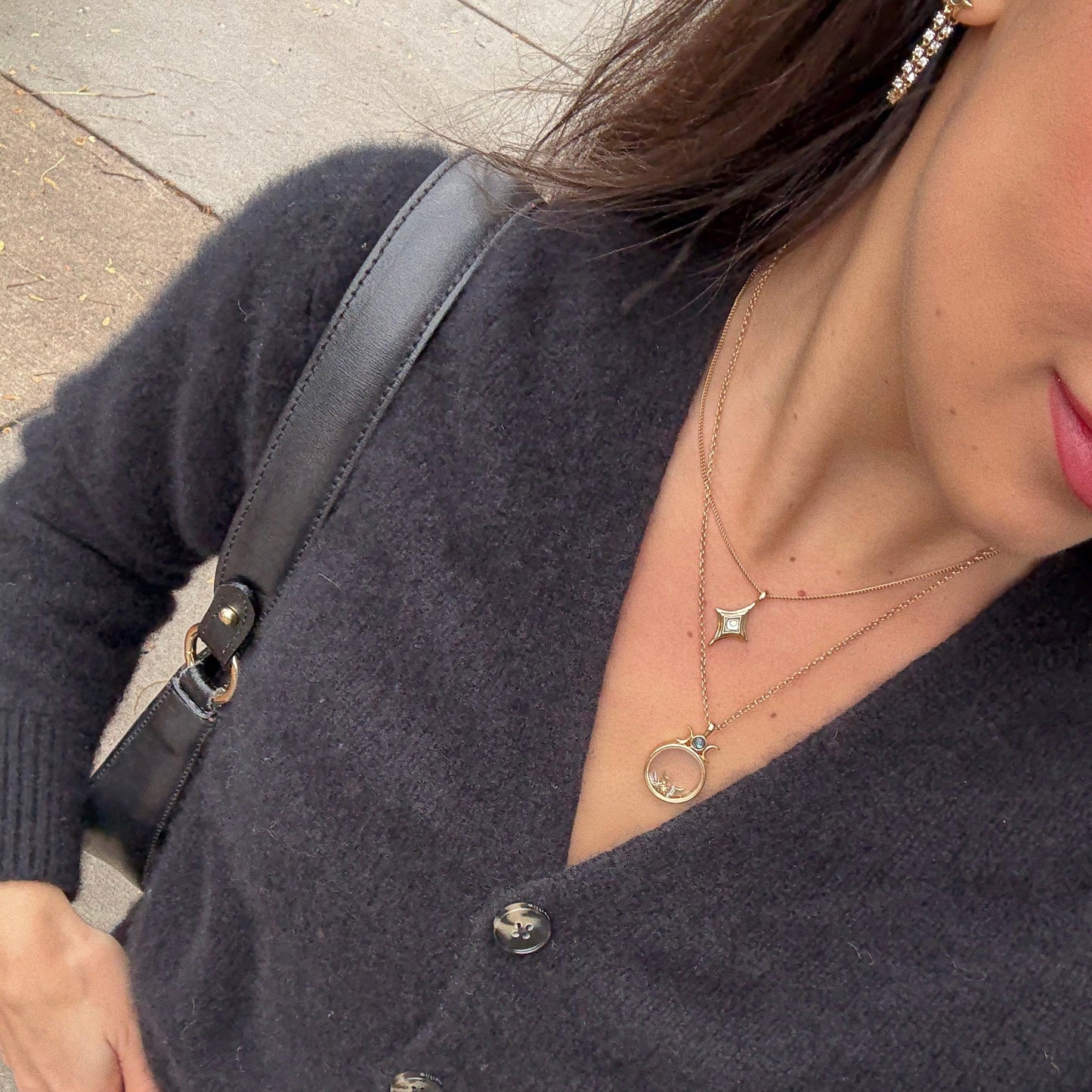 A woman is seen from the shoulders down outdoors, wearing a black fuzzy cardigan, black shoulder bag, layered gold necklaces—including the Wishkeeper Floating Locket Necklace—and a dangling earring.