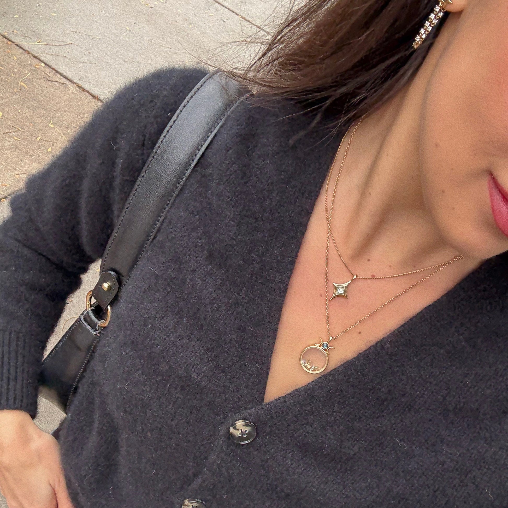 A woman is seen from the shoulders down outdoors, wearing a black fuzzy cardigan, black shoulder bag, layered gold necklaces—including the Wishkeeper Floating Locket Necklace—and a dangling earring.