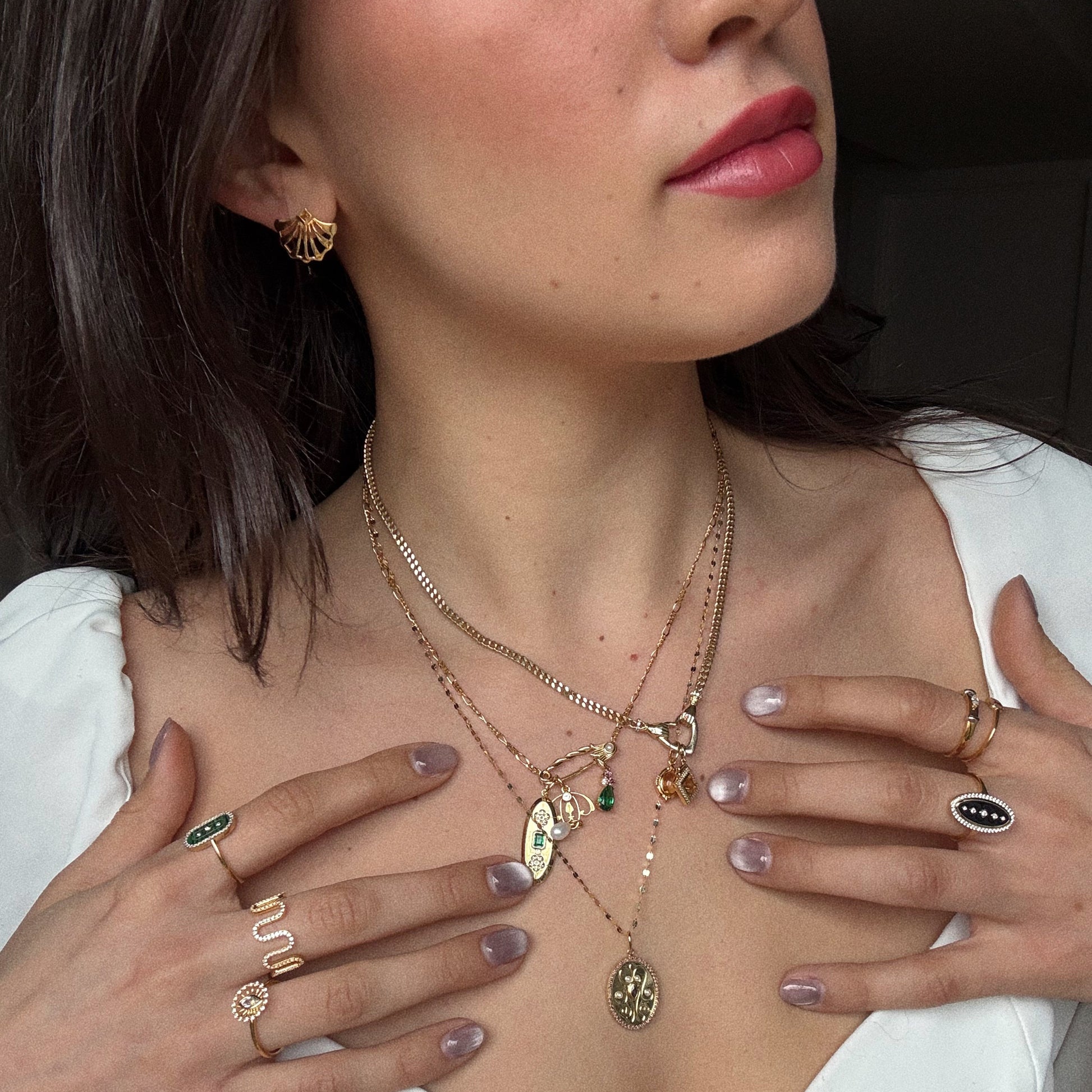 A woman in a white blouse touches her chest, showing manicured nails with a glossy finish while wearing layered gold necklaces, including the Vintage Inspired Pendant Charm, along with gold rings and earrings.