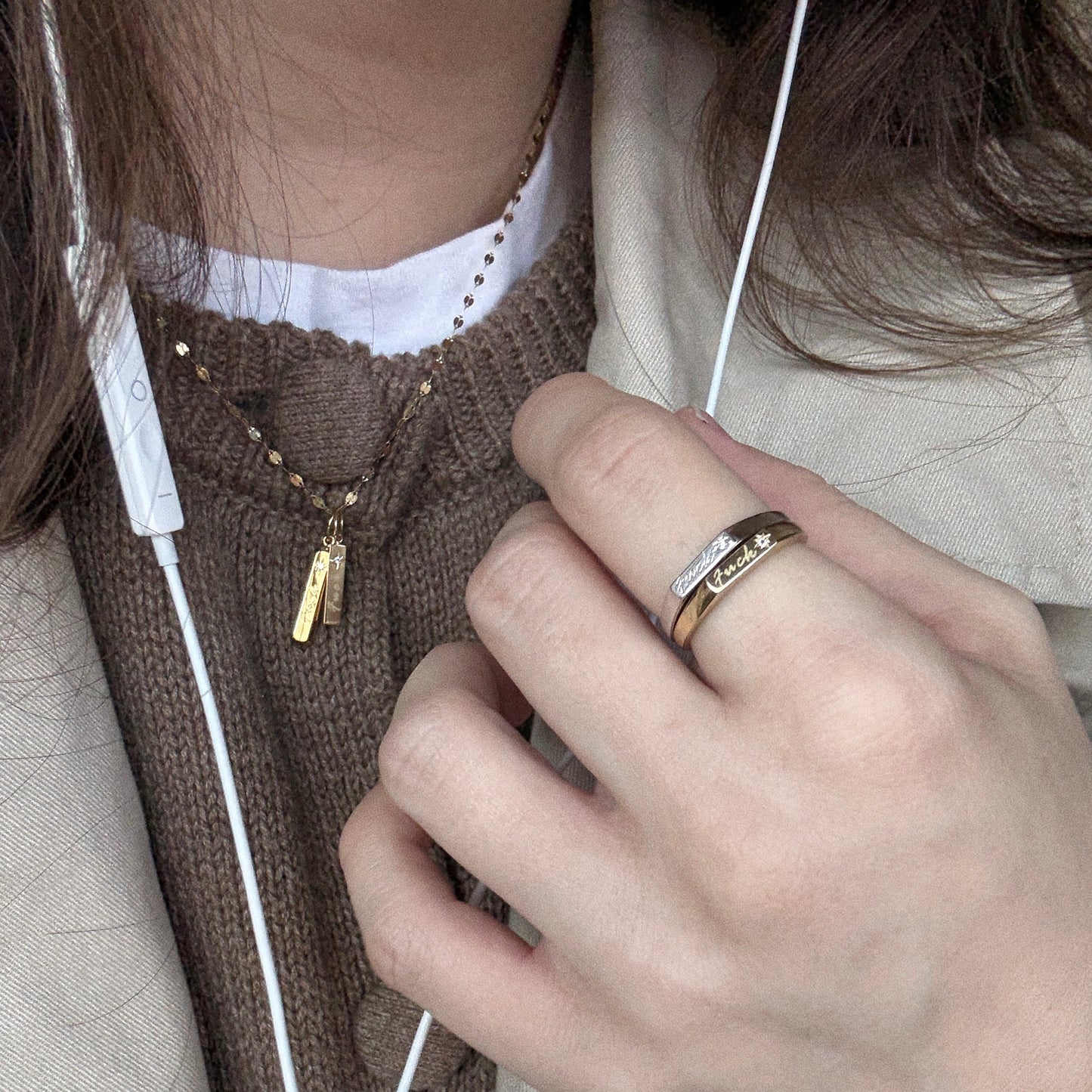 A person in a brown sweater and beige coat holds their hand to their chest, showing off the 14k gold plated Tiny Fuck Gift Set ring and layered necklaces. White earphones dangle from their ears, emphasizing this standout jewelry gift set.