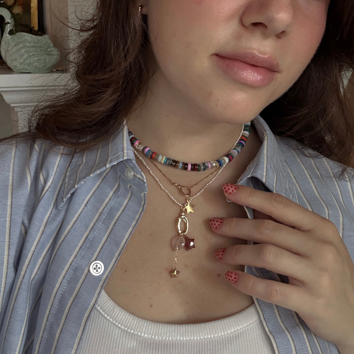 A woman in a striped shirt and white tank top displays her Solid Gold Statement Charm necklace, holding her hand to her chest to show pink nails with black polka dots. Her face is partially visible.