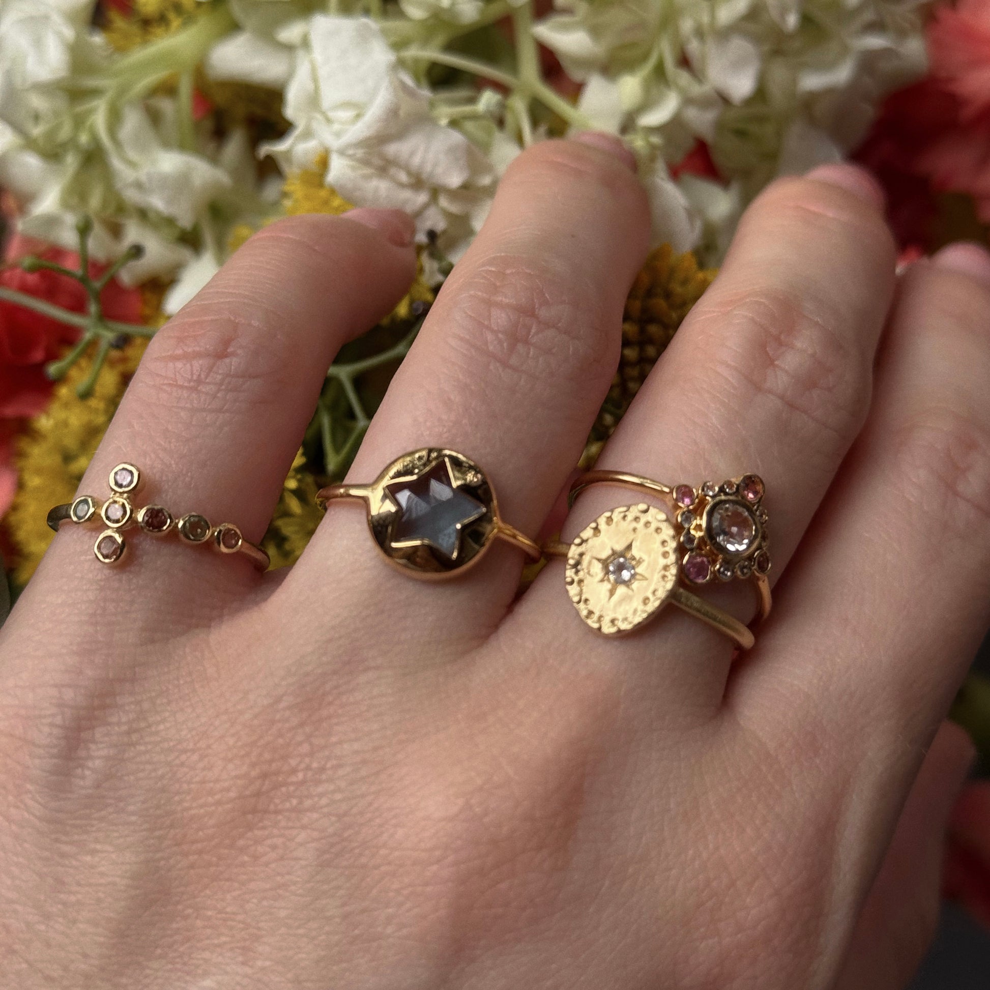 A hand wearing three gold rings, featuring the Brahma Ring in yellow gold plate and a white topaz ring, is held before a vibrant backdrop of blurred white, yellow, and red flowers.
