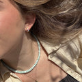 A close-up of a person wearing checkered huggie earrings, a cream-colored shirt, and a turquoise beaded necklace, with sunlight highlighting their light brown hair and skin.