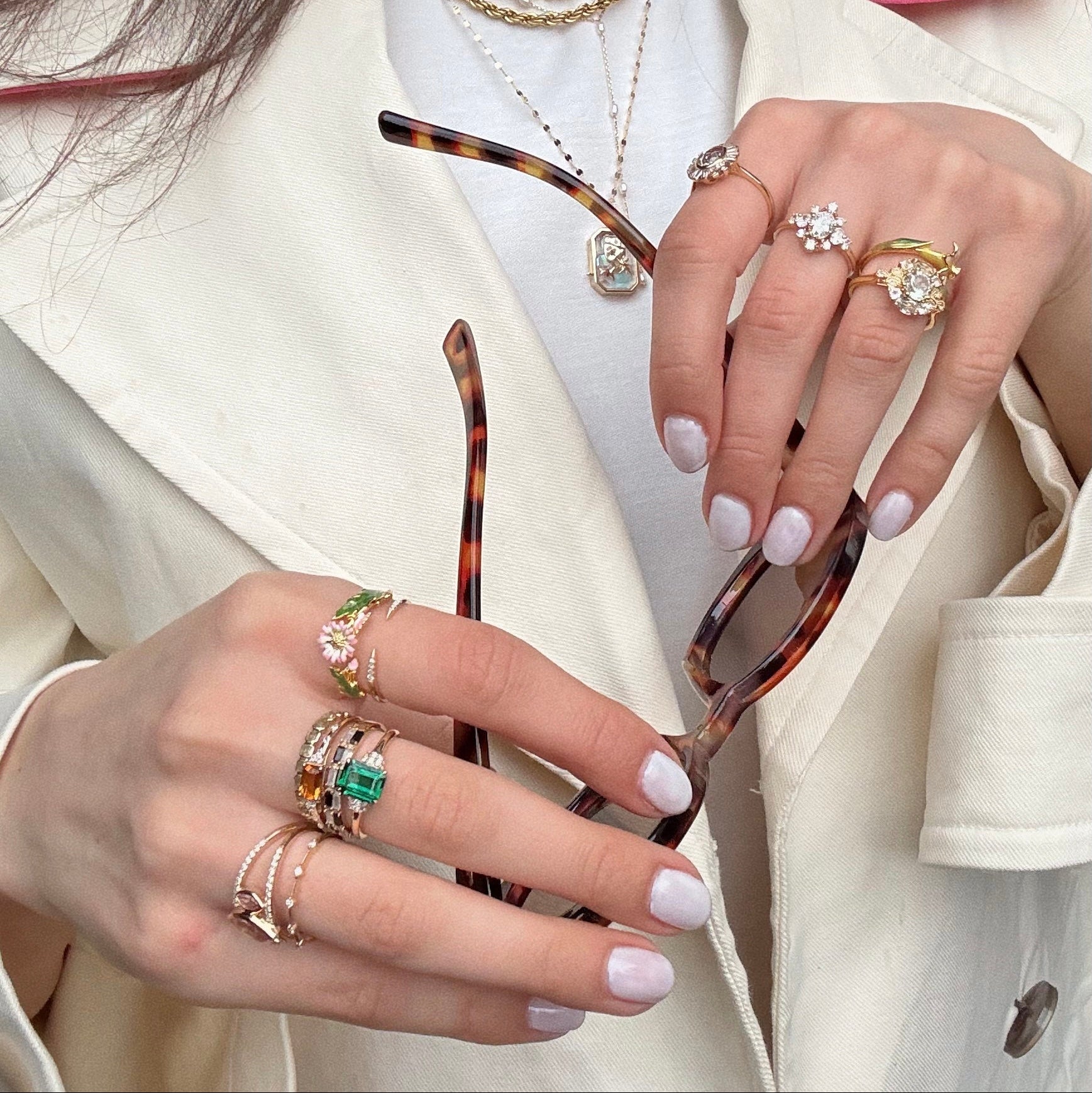 A person in a cream blazer and white top holds tortoiseshell sunglasses. Their hands show the 14k Floating Deco Cluster Diamond Ring, featuring diamonds set in solid yellow gold, paired with layered necklaces for a Local Eclectic exclusive vibe.