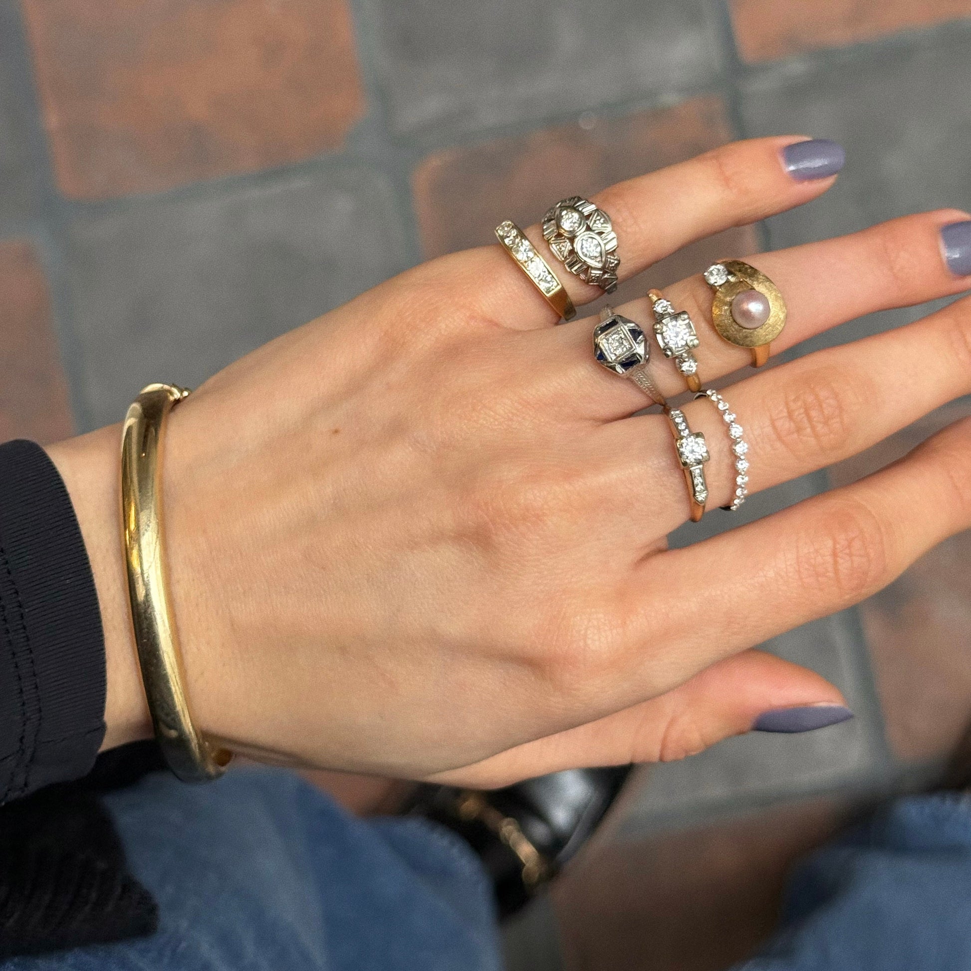 A hand with light purple nails displays several gold and silver gemstone rings and the Vintage 9k Classic Bangle against a tiled floor background.