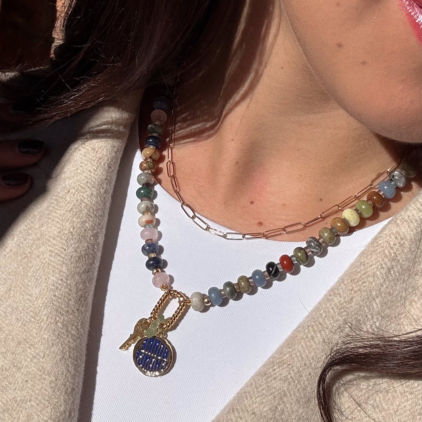 Wearing a beige coat and white top, a woman showcases the Macey Customizable Beaded Charm Holder Necklace with two round pendants, paired with a thin gold chain. Sunlight highlights her wavy brown hair and jewelry.