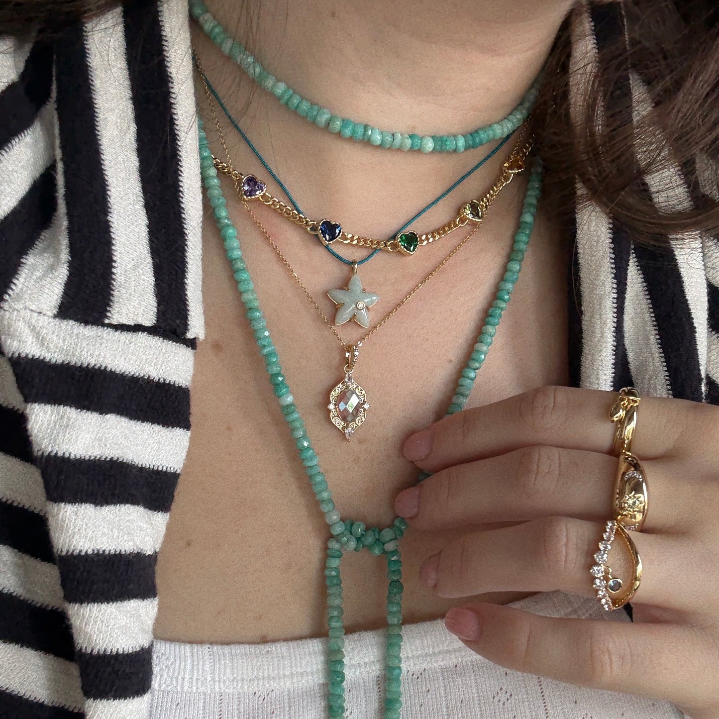 A person wearing layered turquoise beaded necklaces, the Lucky Star Amazonite Pendant Necklace (14k gold plated), and gemstone gold rings, dressed in a black and white striped jacket.
