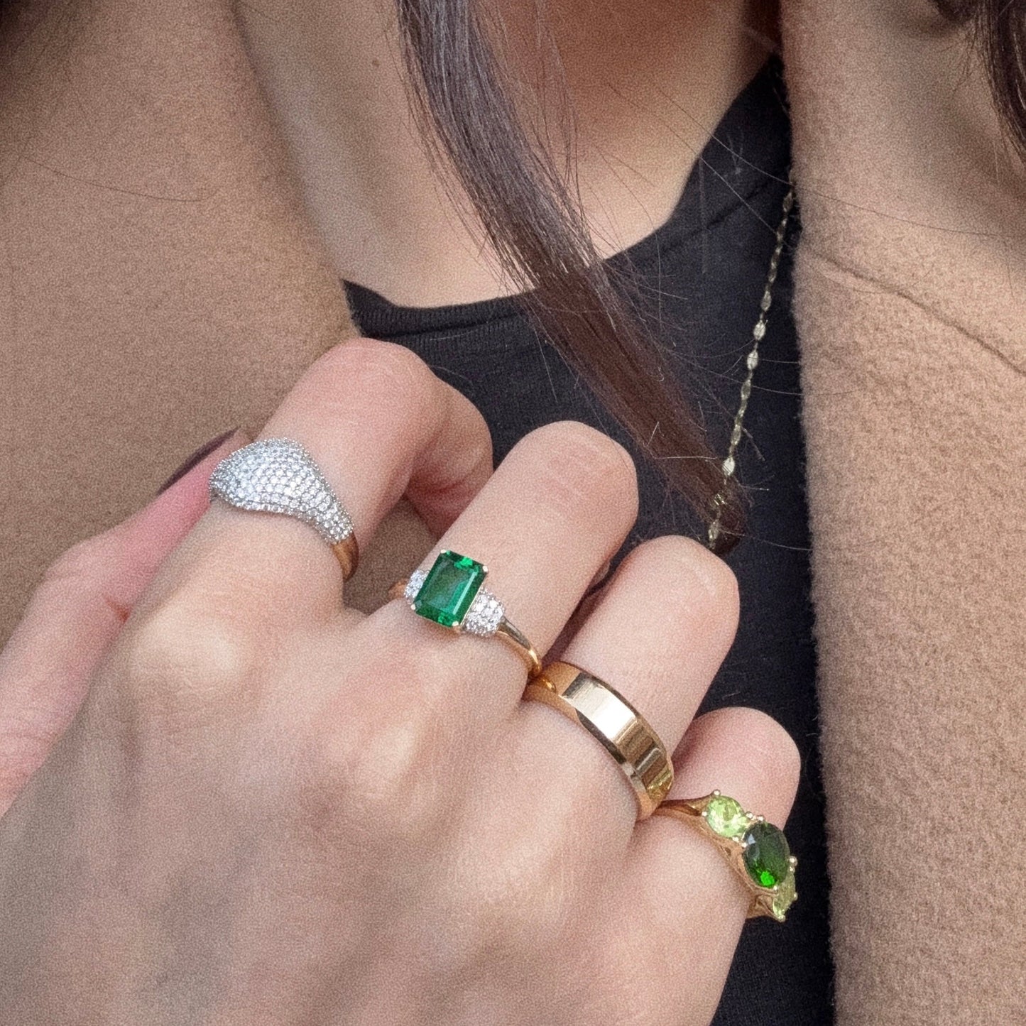 A close-up of a hand in a black top and beige coat showcases four rings, including the 14k Solid Gold Pave Diamond Signet Ring (sample size 7), a green gemstone ring, a gold band, and one with two small green stones.