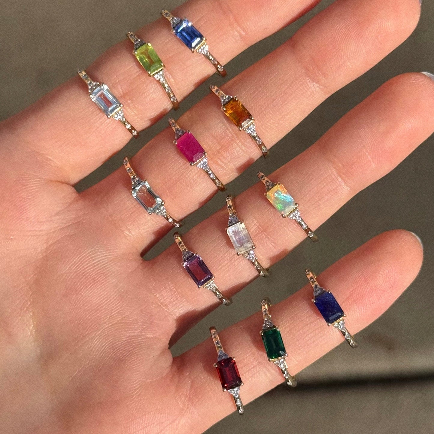 A hand shows twelve gold rings, including the Solid Gold Glimmering Birthstone Baguette Ring, each featuring vibrant rectangular stones and arranged neatly along the fingers.