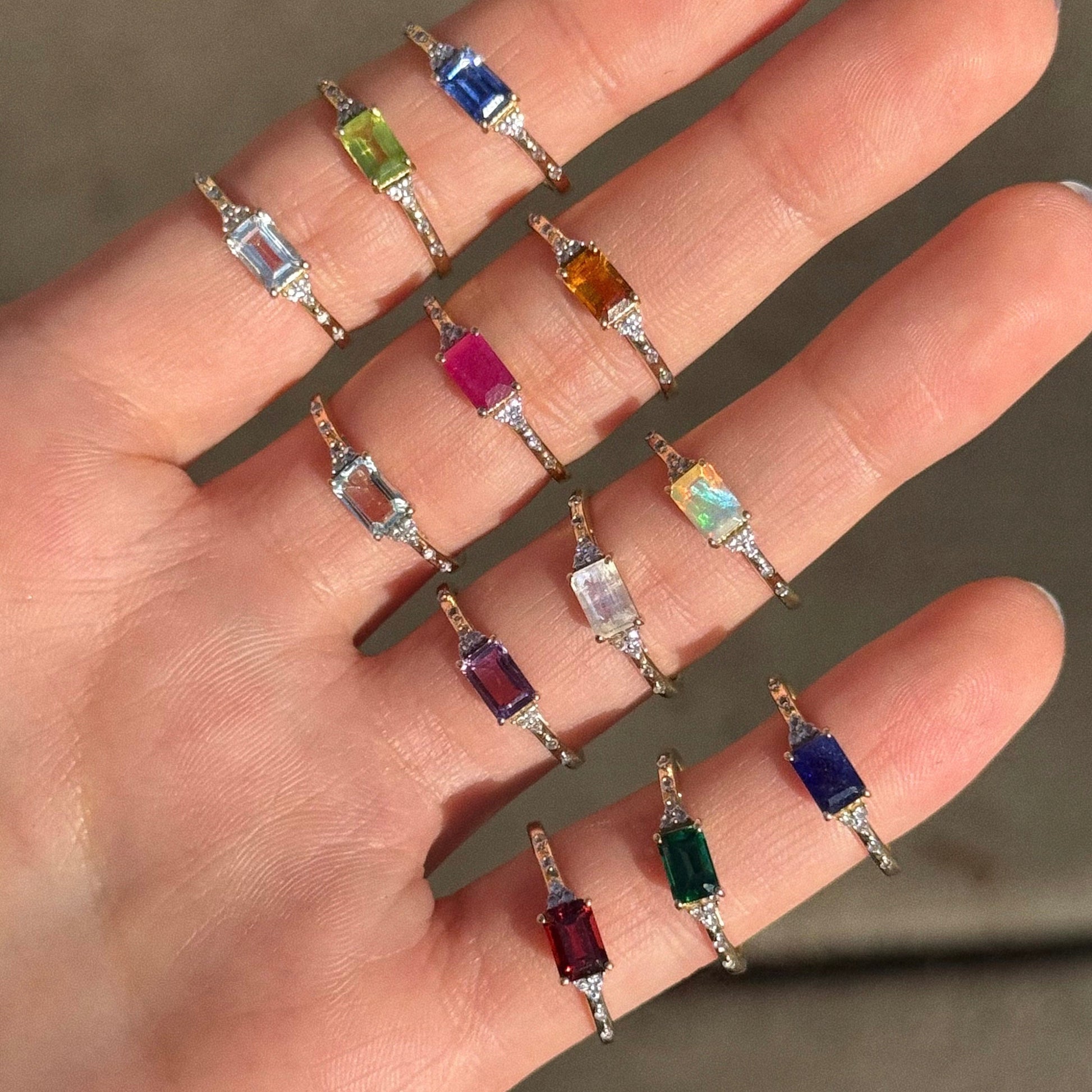 A hand shows twelve gold rings, including the Solid Gold Glimmering Birthstone Baguette Ring, each featuring vibrant rectangular stones and arranged neatly along the fingers.