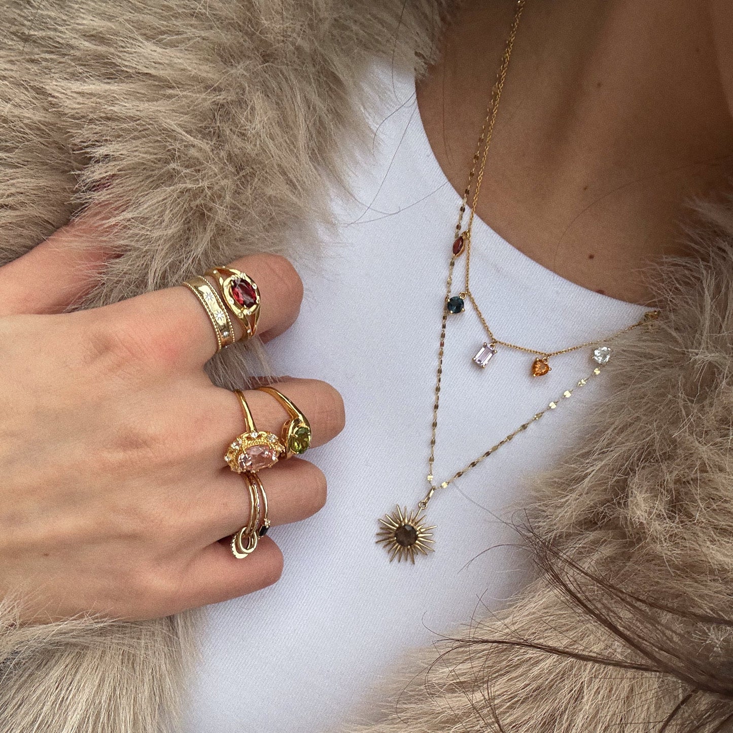 A person in a white top and faux fur coat layers gold necklaces—a sunburst pendant and the Classical Confetti Gemstone Necklace with charms—plus multiple 14k gold vermeil rings with multicolored gemstones.