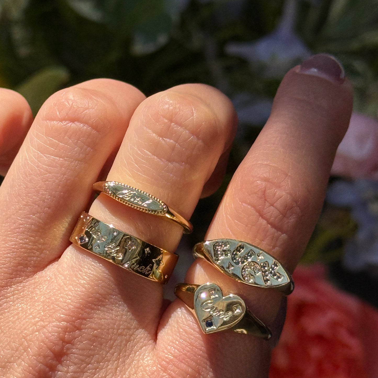 A hand gleams in sunlight, wearing four Colorful Language Fuck Signet Rings in 14k yellow gold—each one uniquely engraved and designed—set against blurred flowers for a bold, colorful statement.